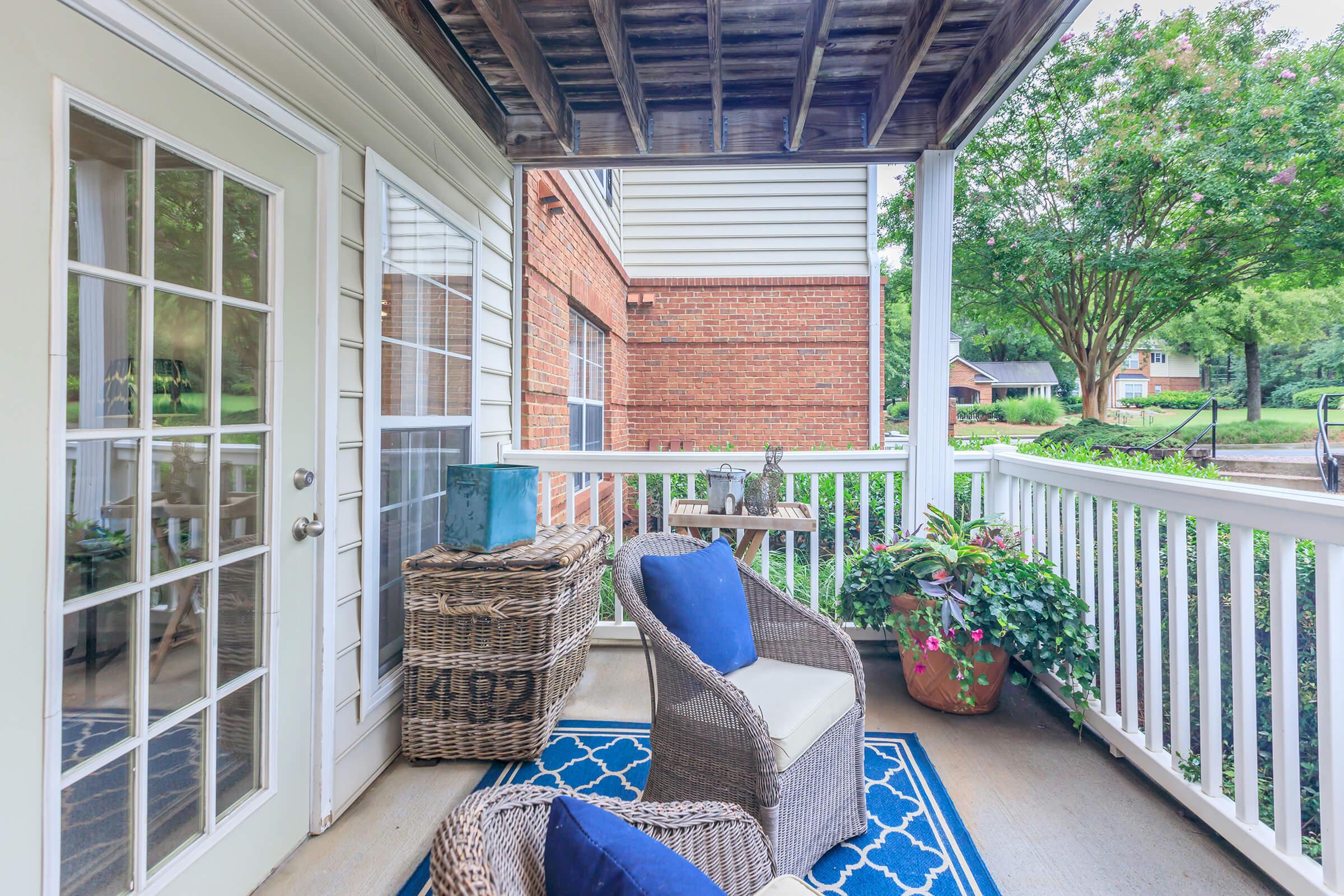 A cozy outdoor balcony featuring a wicker chair with blue cushions, a small wooden table, and decorative plants. The area is well-lit with natural light, surrounded by a white railing and trees in the background, creating a peaceful atmosphere.