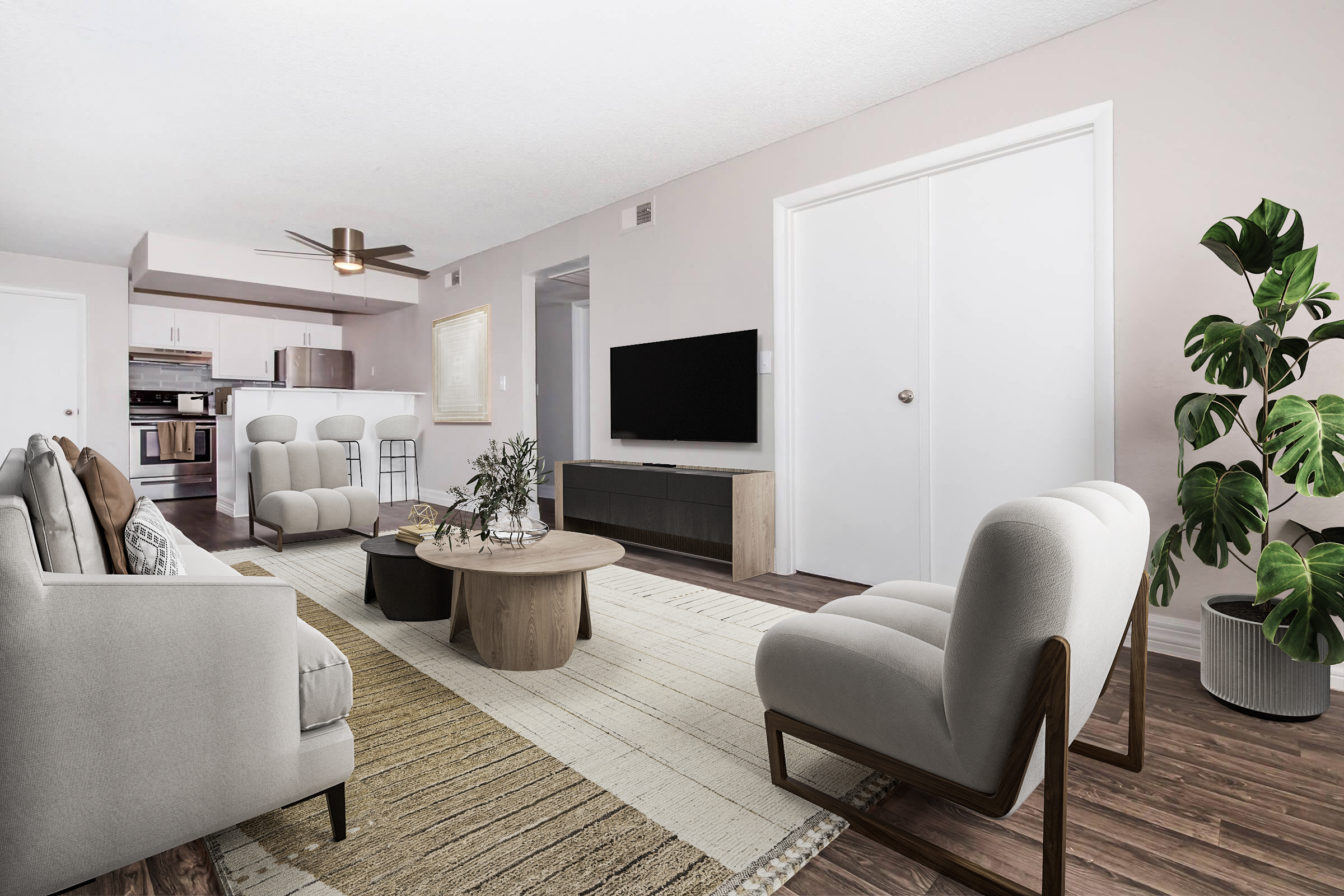 A modern living room featuring light-colored walls and hardwood flooring. The space includes a comfortable seating area with soft gray armchairs and a sofa, a round wooden coffee table, a decorative plant, and a TV unit. A ceiling fan is visible, along with a partially open door leading to another room.