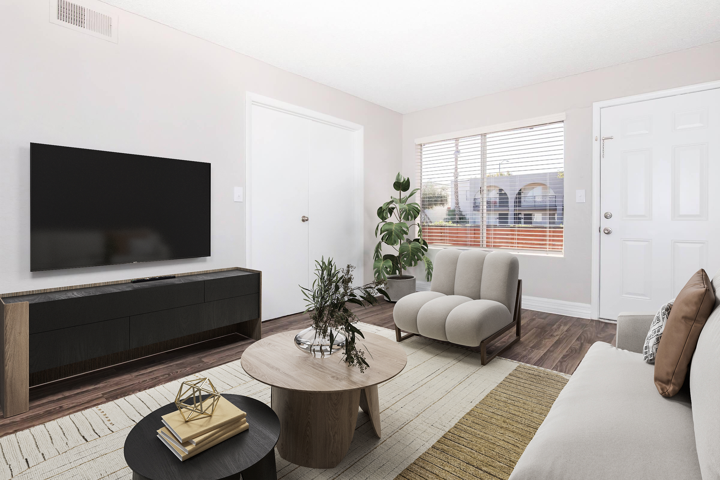 A modern living room featuring a large flat-screen TV mounted on a wall, a stylish coffee table, a light-colored sofa, and a pair of contemporary armchairs. A potted plant adds greenery, and a window with blinds allows natural light in, illuminating the space with a cozy atmosphere.
