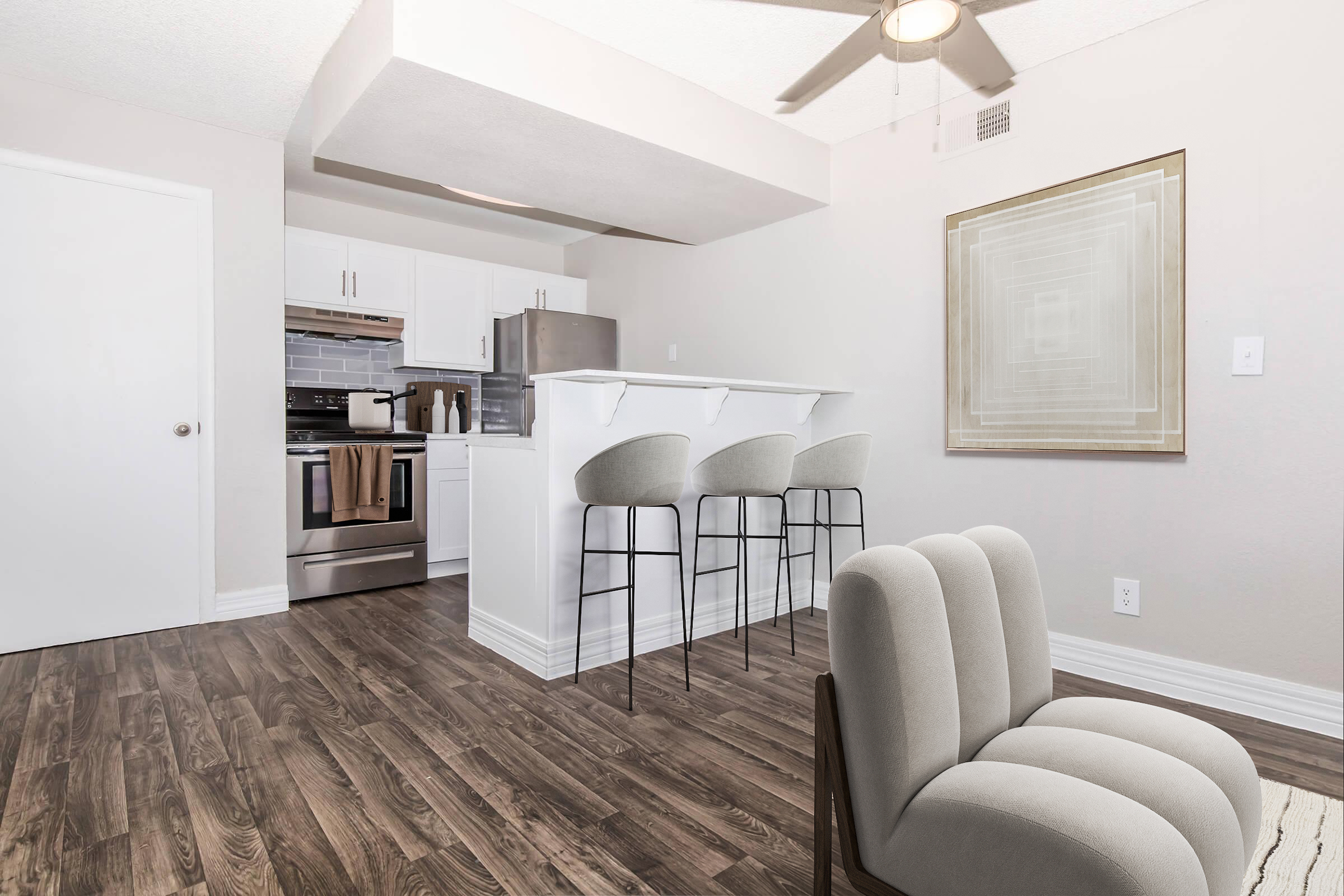 A modern kitchen with white cabinetry, stainless steel appliances, and a bar area featuring three high stools. The open space has wooden flooring and a neutral-colored wall with a geometric art piece. A comfortable, beige armchair is positioned nearby, adding a stylish touch to the room.