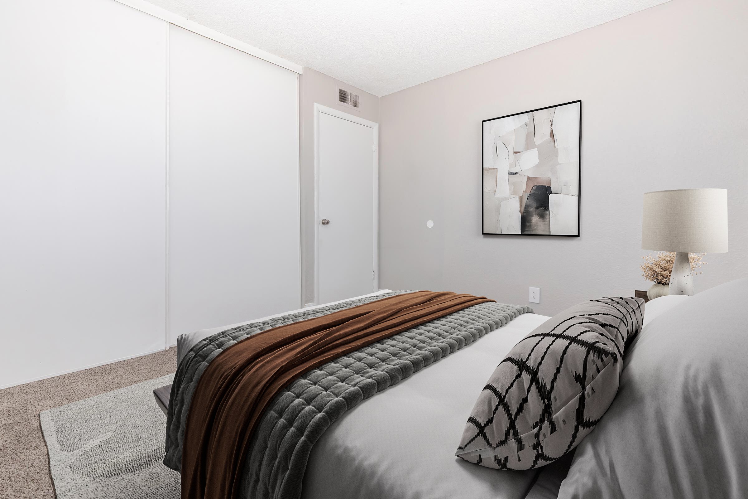 A cozy bedroom featuring a neatly made bed with gray and brown bedding, a decorative pillow, and a soft rug on the carpeted floor. There are light gray walls, a piece of abstract wall art, and a lamp on the bedside table. A closet door and a white door are visible in the background.