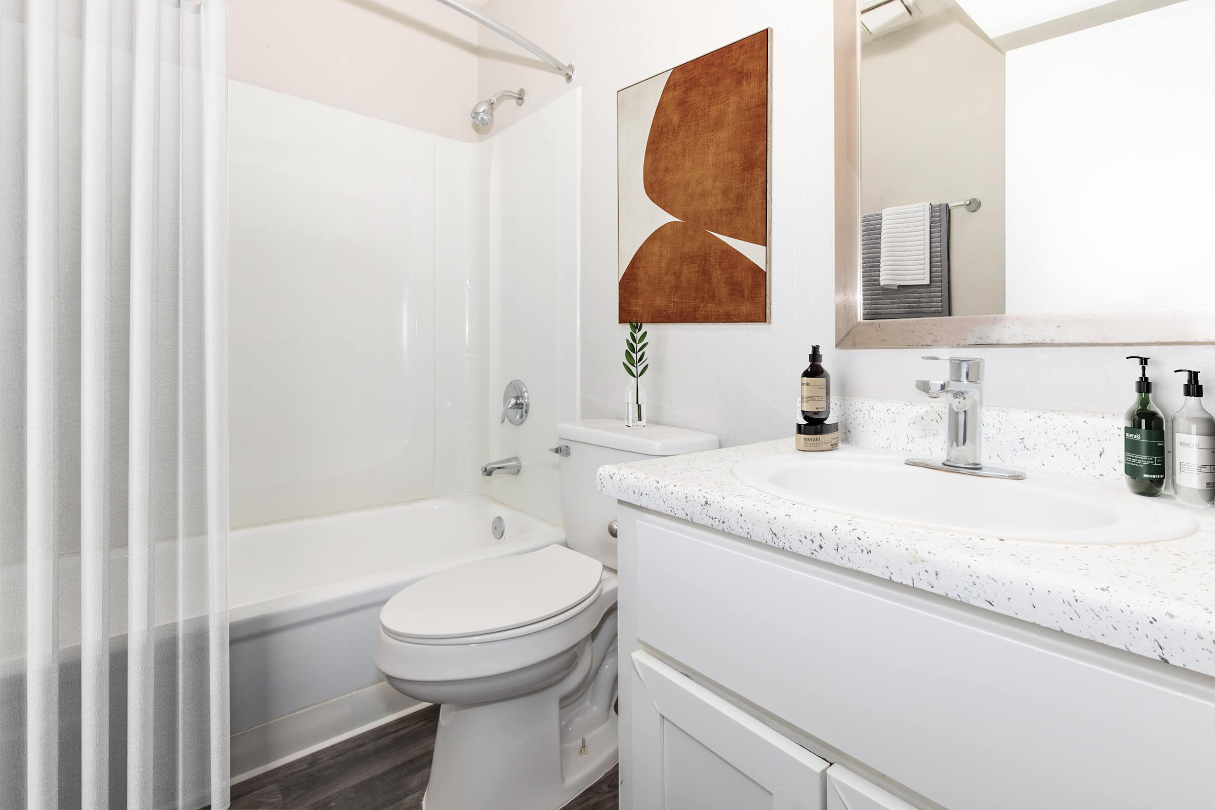 A clean and modern bathroom featuring a bathtub with a shower curtain, a white vanity with a sink, and decorative items including a small plant and toiletries. A piece of abstract wall art adds a contemporary touch to the space.