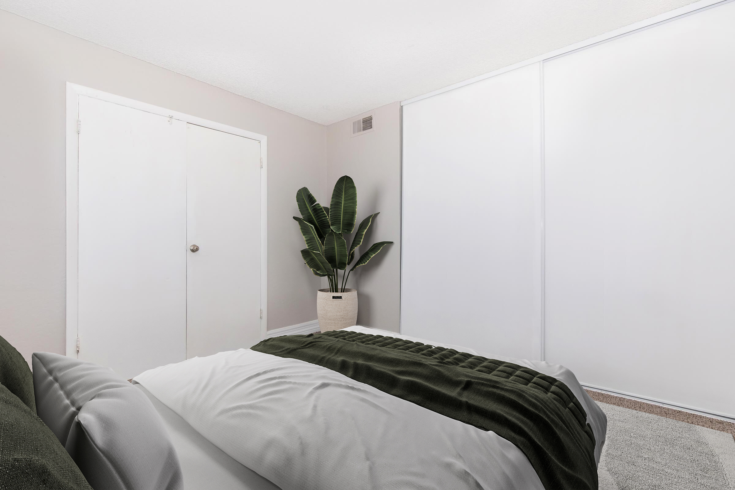 A minimalist bedroom featuring a bed with a gray and dark green comforter, a large potted plant in the corner, and a closet door. The walls are light beige, and there are large white sliding doors, creating a clean and serene atmosphere.