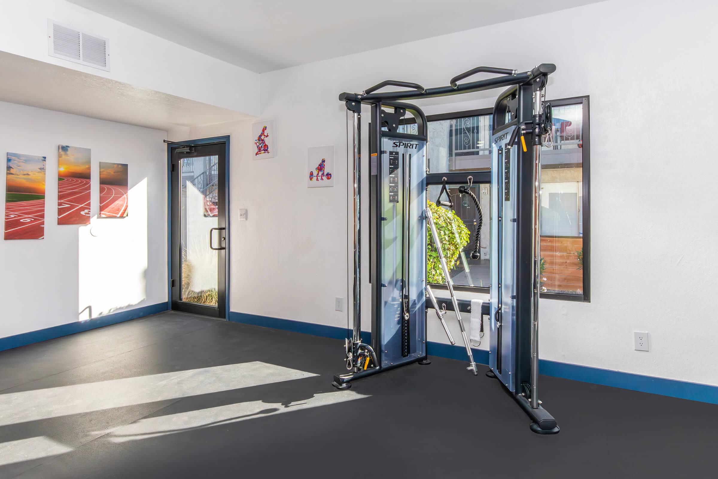 A modern workout space featuring a cable machine against a bright wall. The room has a large window with natural light streaming in and is decorated with sports-themed artwork. The floor is covered in dark rubber material, providing a fitness-friendly environment.