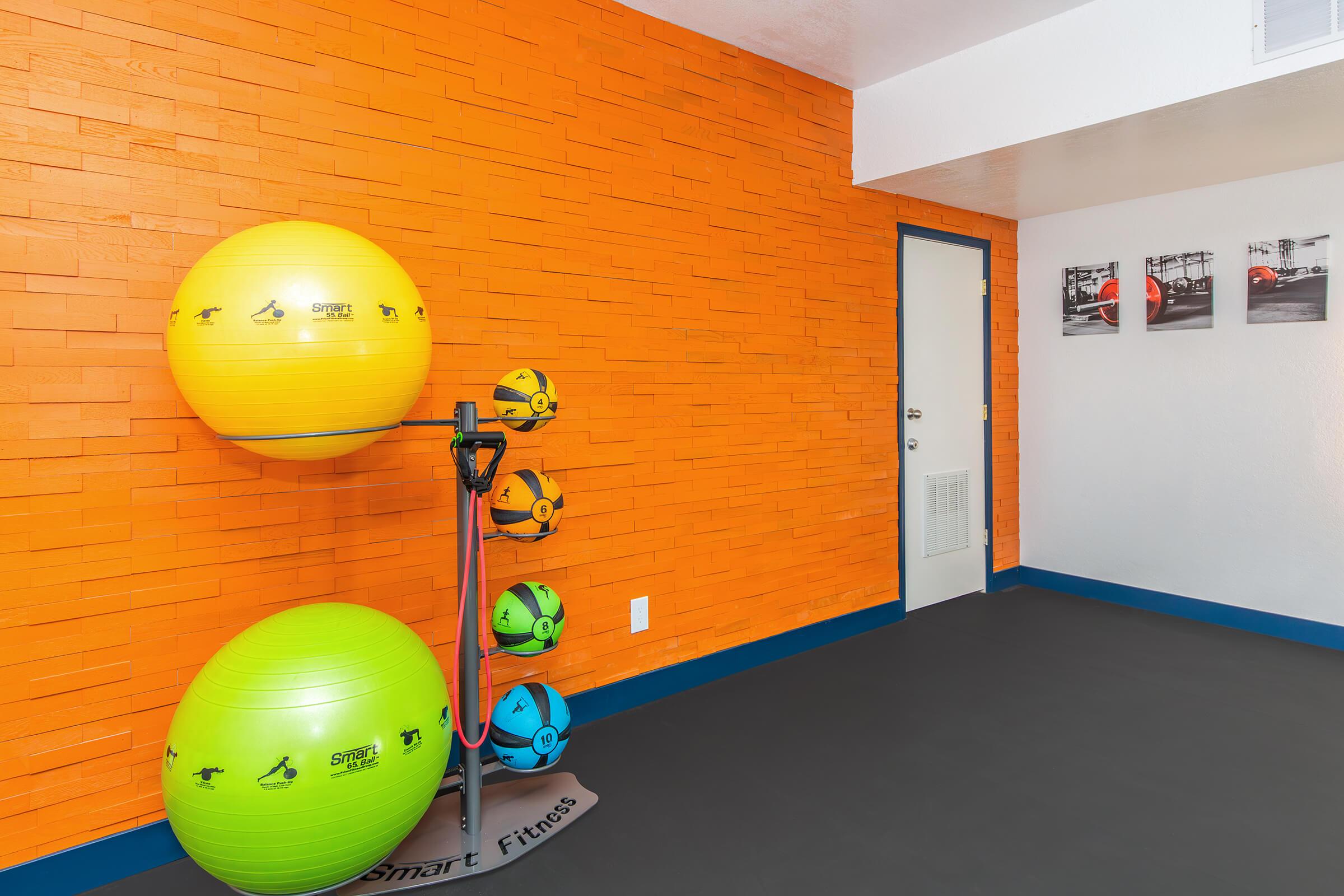 Interior of a fitness room featuring an orange accent wall. There are two large exercise balls in yellow and green stacked on a holder, and smaller colorful balls displayed on a rack. A door is visible on one side, and three framed black-and-white photos are hung on the wall opposite.