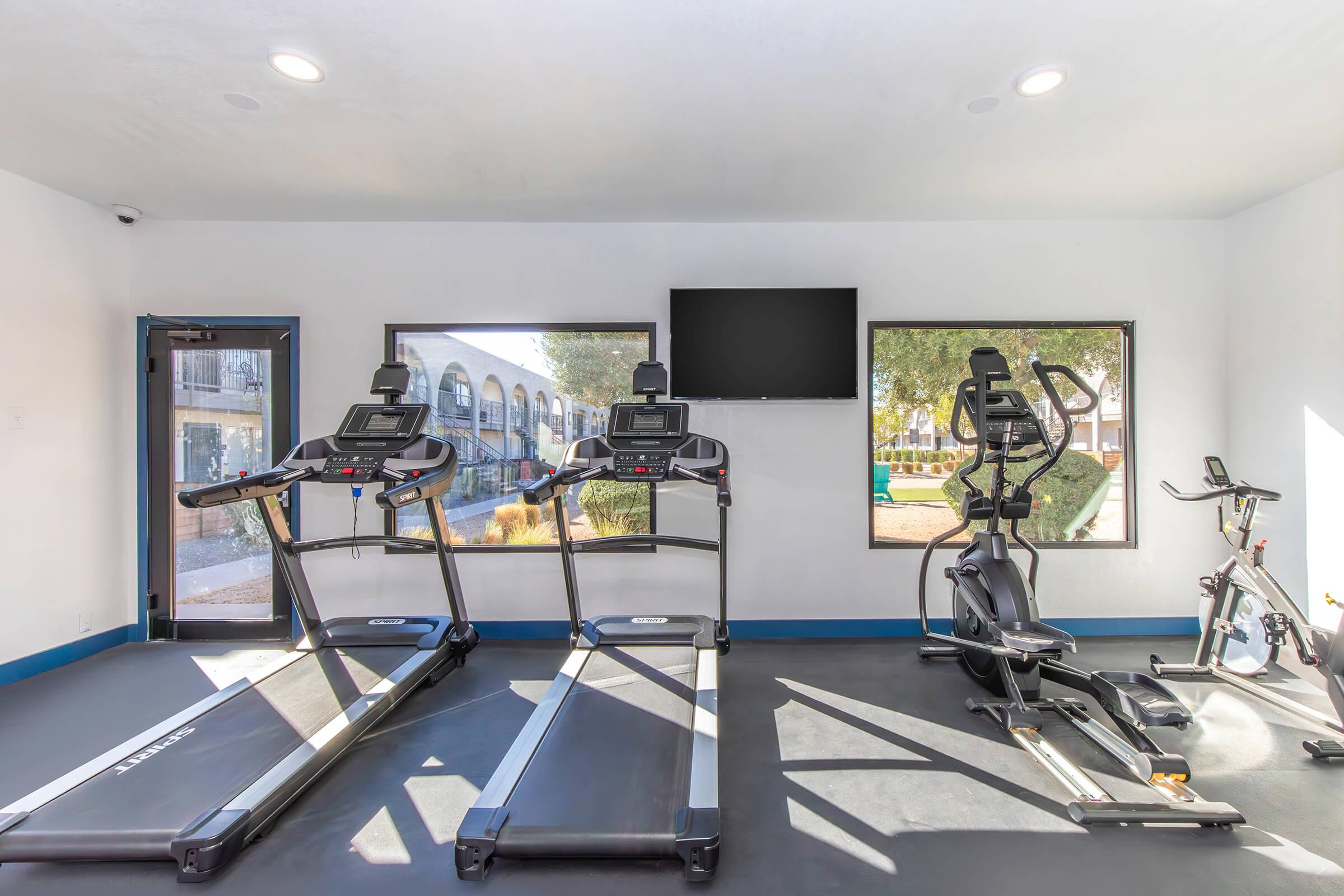 A bright fitness room featuring two treadmills and an elliptical machine, all positioned on a dark rubber floor. Large windows allow natural light, and a flat-screen TV is mounted on the wall, enhancing the workout environment. The space appears clean and inviting, with a view of a landscaped area outside.