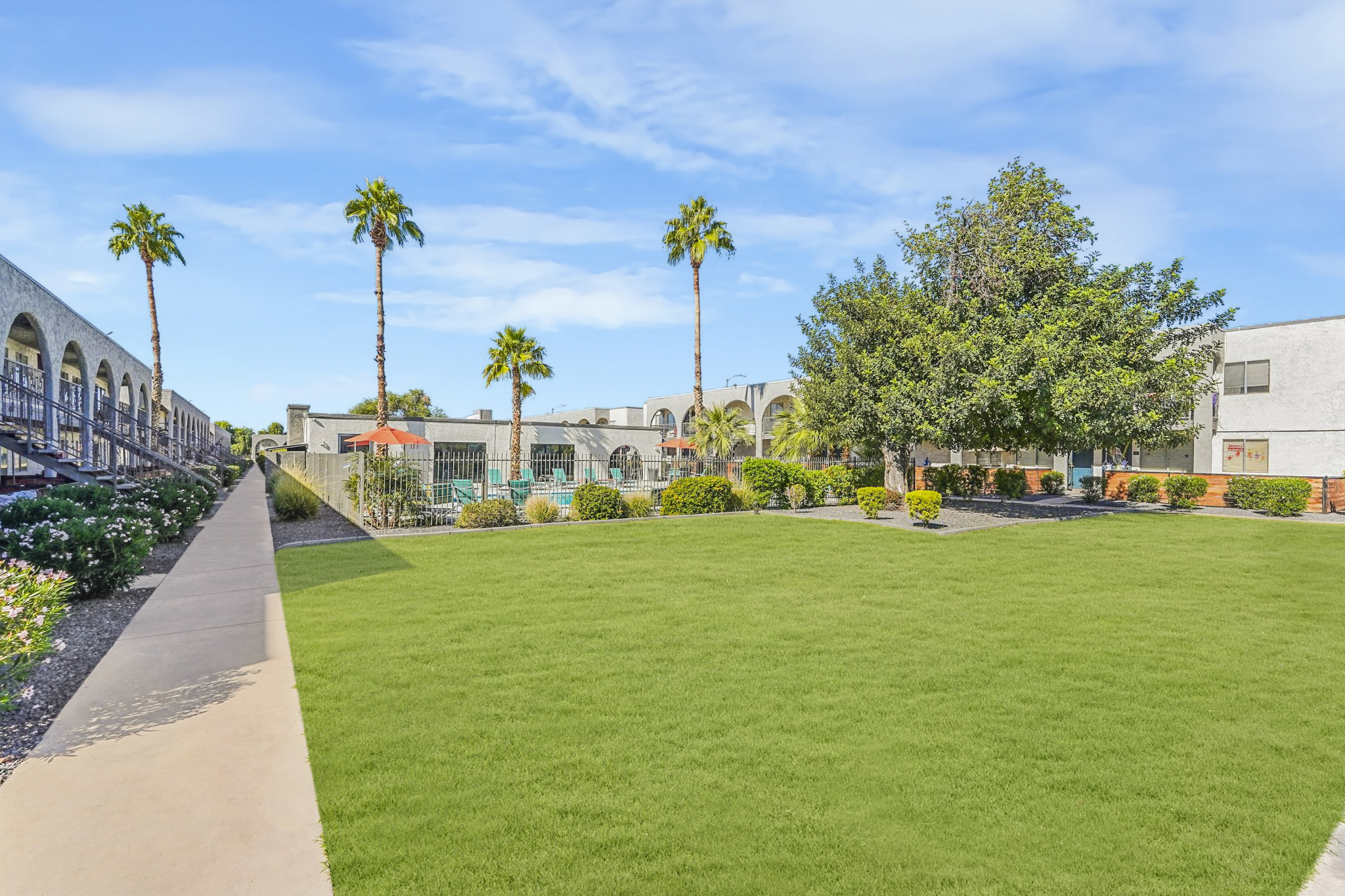 A landscaped courtyard featuring a well-maintained grassy area, palm trees, and shrubs. Surrounding the space are multi-story buildings. A swimming pool with lounge chairs is partially visible in the background, along with an umbrella-covered sitting area. The sky is clear and blue, creating a serene environment.