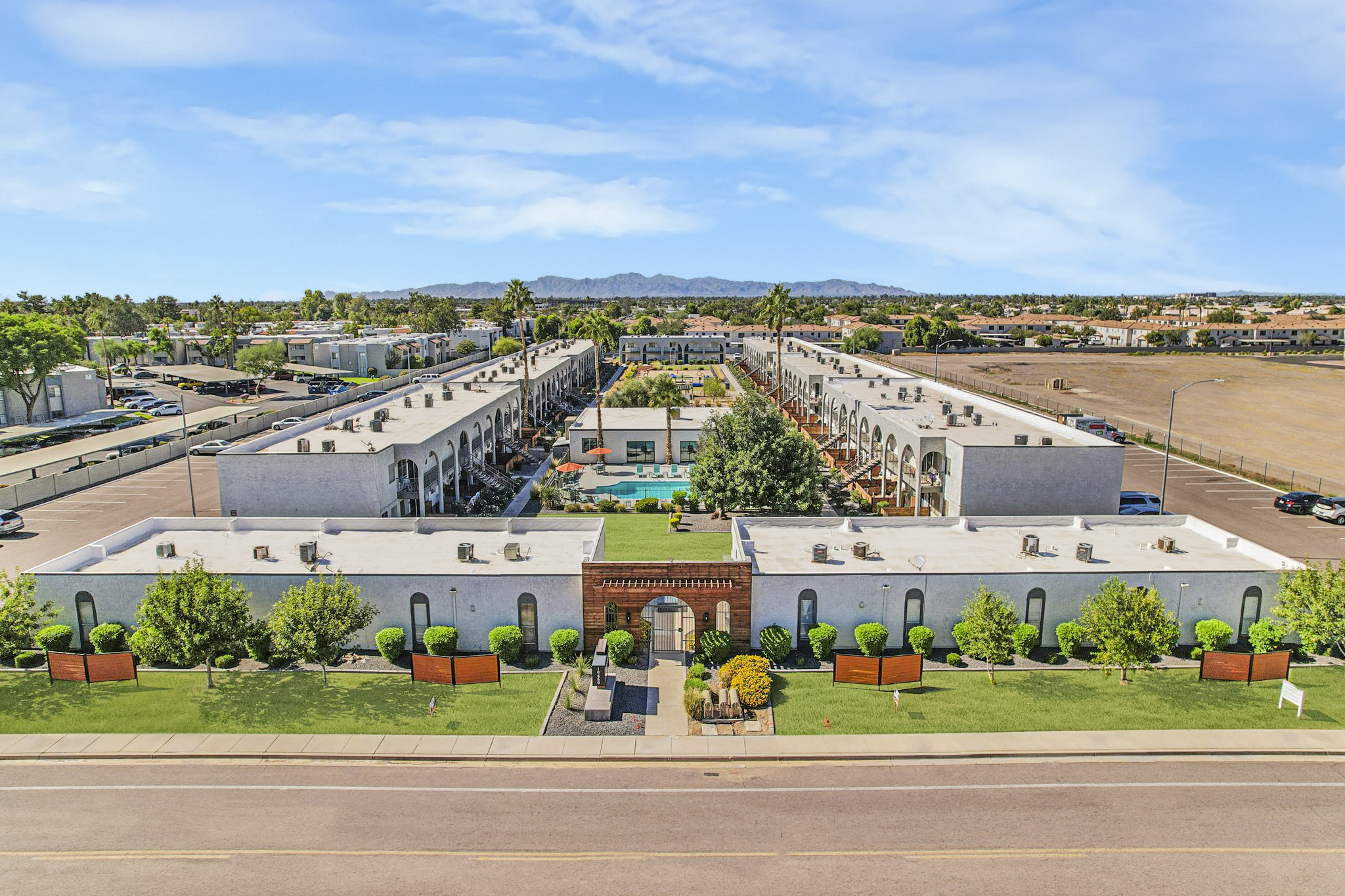 Aerial view of a landscaped residential complex featuring multiple buildings arranged around a central pool area. The setting includes well-maintained greenery, palm trees, and a mountain backdrop, with a pathway leading through the property. The atmosphere is bright and inviting with clear blue skies.