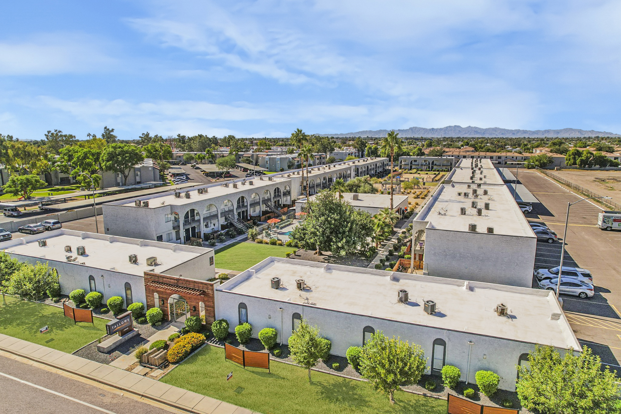 An aerial view of a residential complex featuring multiple buildings with white exteriors, green landscaping, and courtyard areas. The scene includes paved pathways, palm trees, and a mountain backdrop under a clear blue sky.