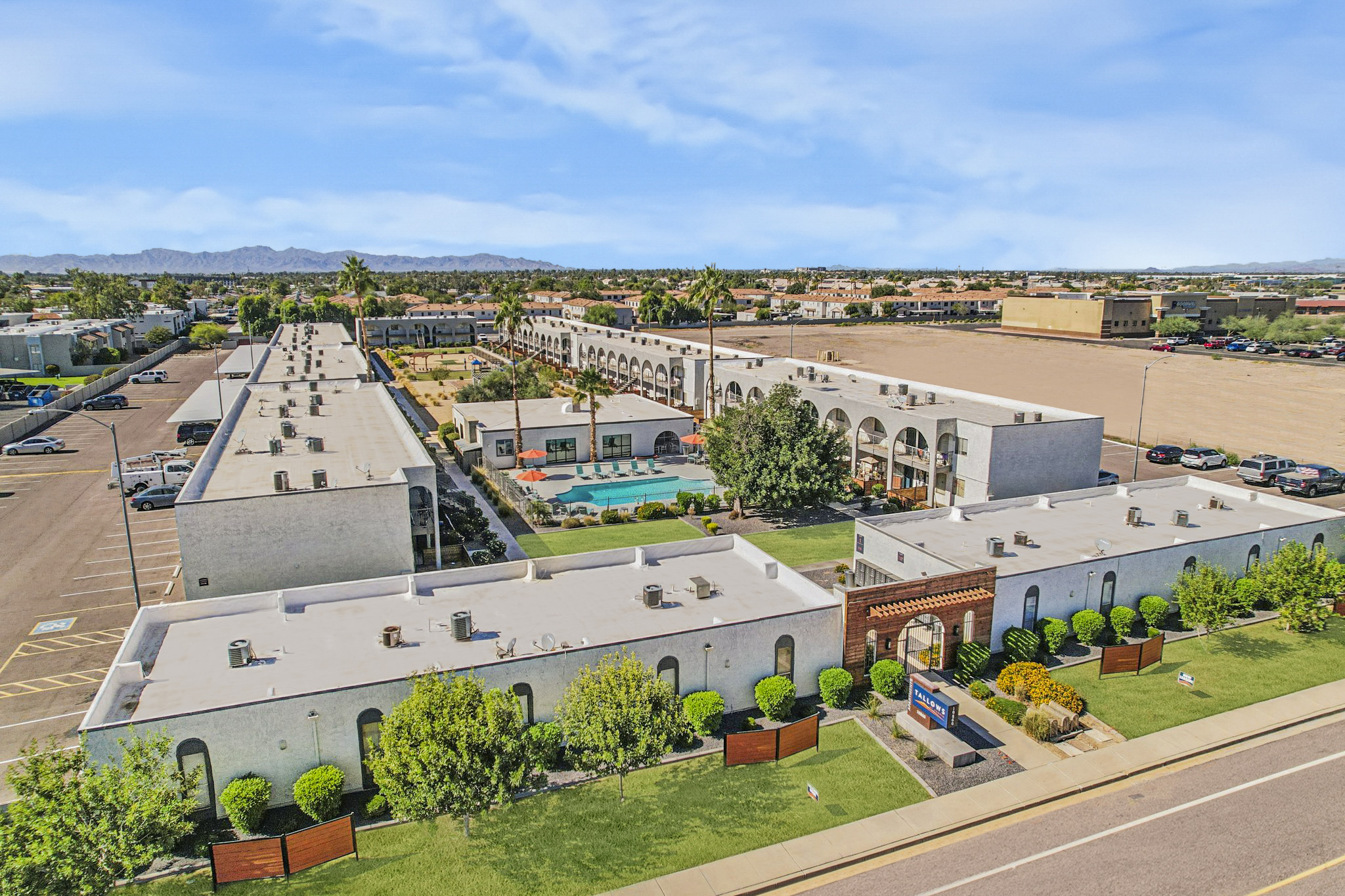 Aerial view of a hotel complex featuring multiple low-rise buildings, a landscaped courtyard with a swimming pool and lounge area. Surrounding the property are parking spaces, palm trees, and distant mountains under a clear blue sky.