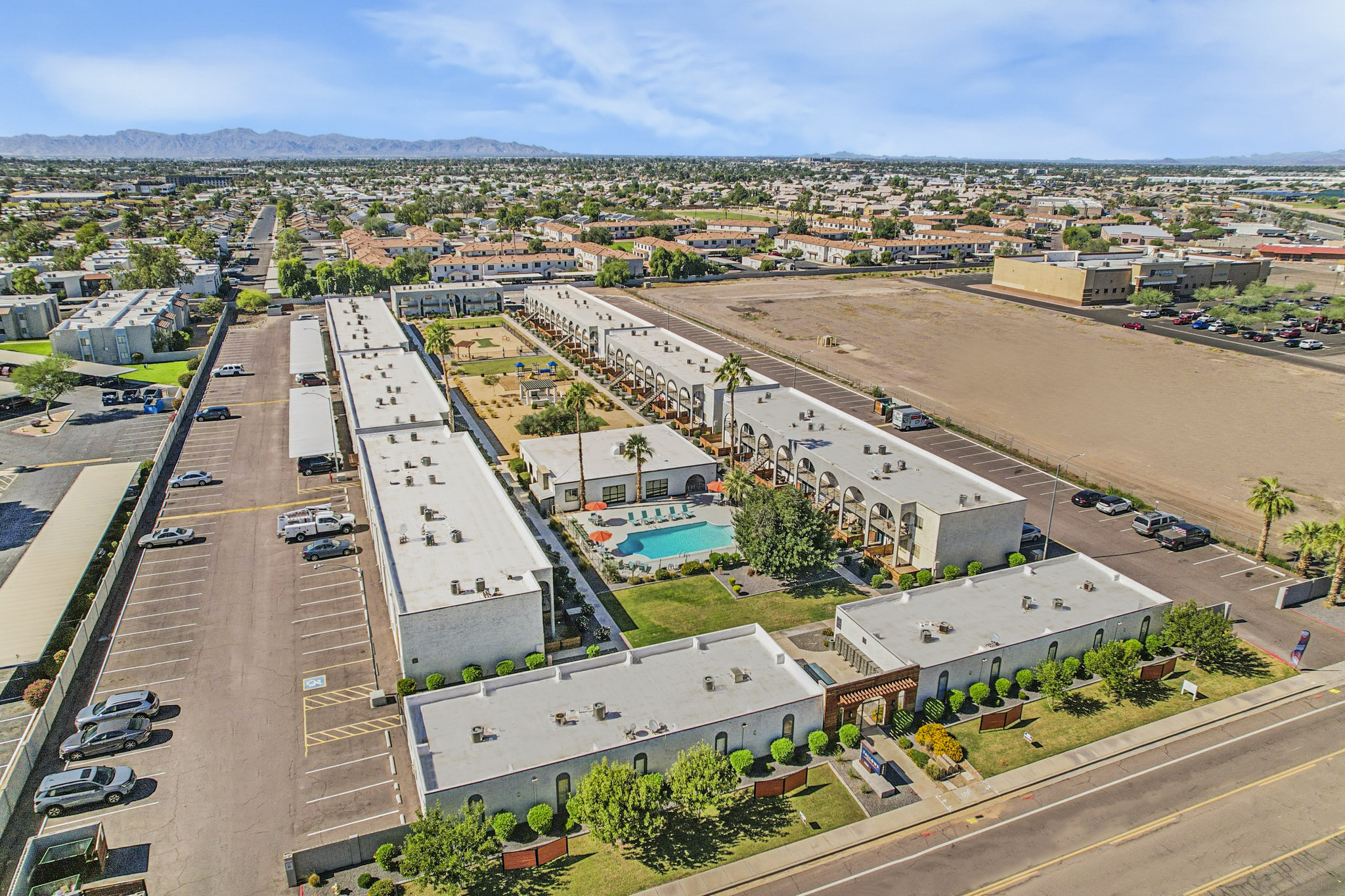 Aerial view of a residential complex featuring multiple two-story buildings with a central swimming pool, surrounded by landscaped areas. The surrounding neighborhood includes additional buildings and open space, with mountains visible in the distance under a clear blue sky. Parking areas can also be seen.