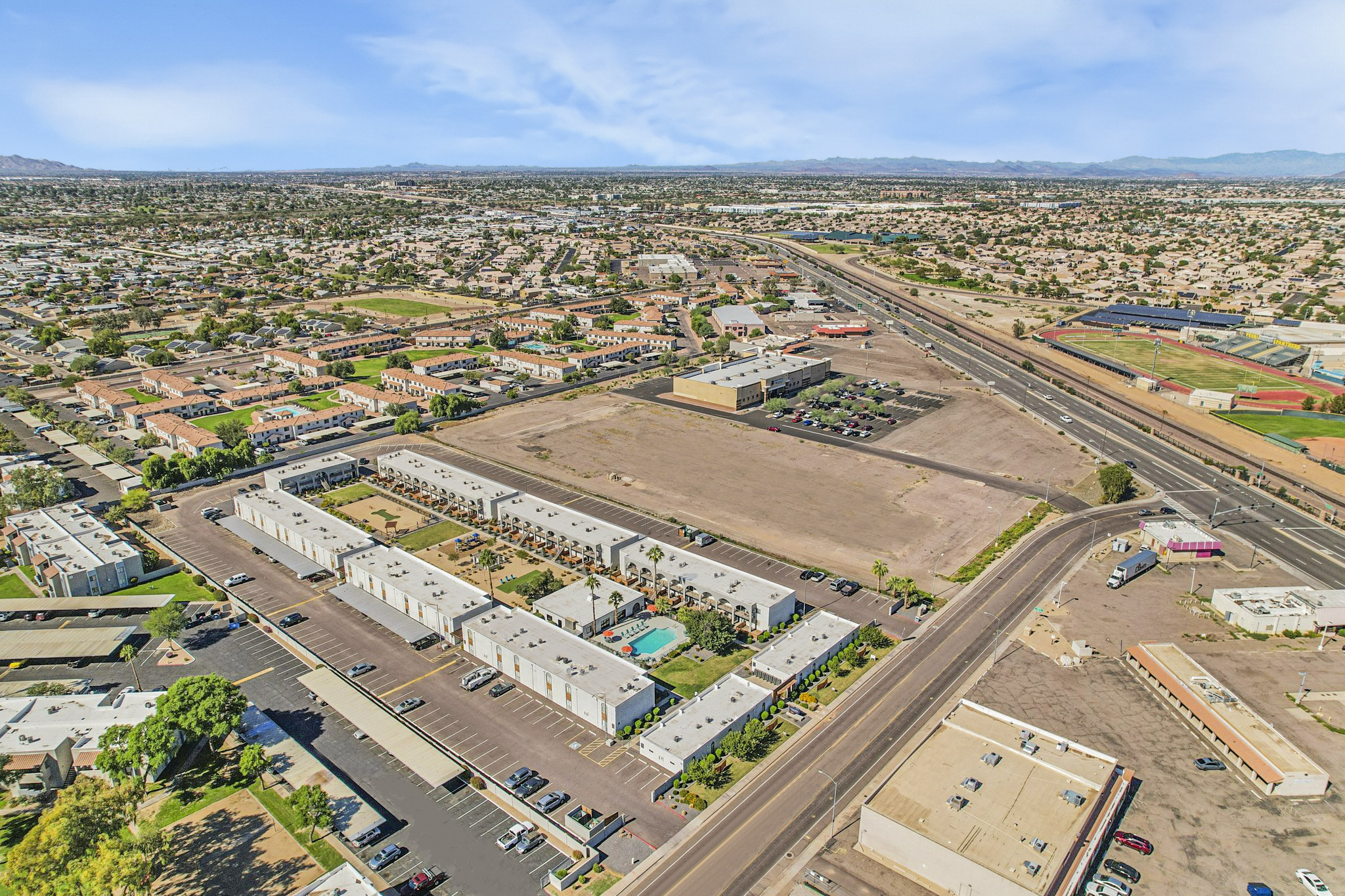 An aerial view of a suburban area featuring low-rise apartment complexes, a vacant lot with a swimming pool visible, and a road lined with parked cars. In the background, there are residential homes and mountains, with railway tracks running parallel to the road. Bright blue skies and scattered clouds are overhead.