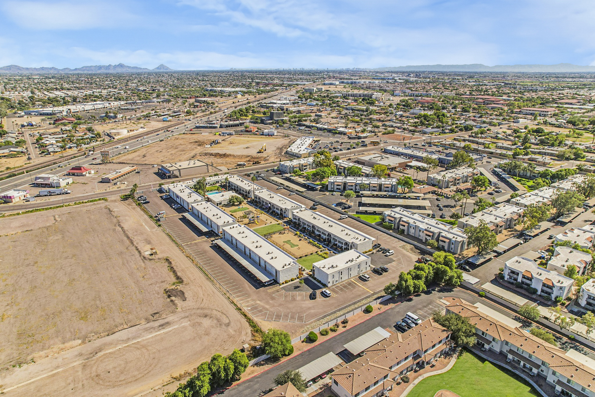 Aerial view of a suburban area featuring multiple apartment complexes, green spaces, and surrounding roads. In the background, there are commercial buildings and open land, with mountains visible in the distance under a clear blue sky.