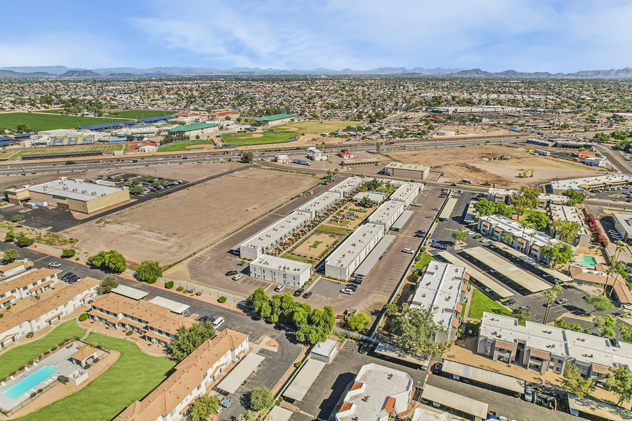 Aerial view of a semi-urban landscape featuring a mix of residential buildings and commercial areas. In the foreground, there are low-rise apartment complexes with pools, while the background shows industrial buildings and agricultural land. Mountains are visible in the distance under a clear blue sky.