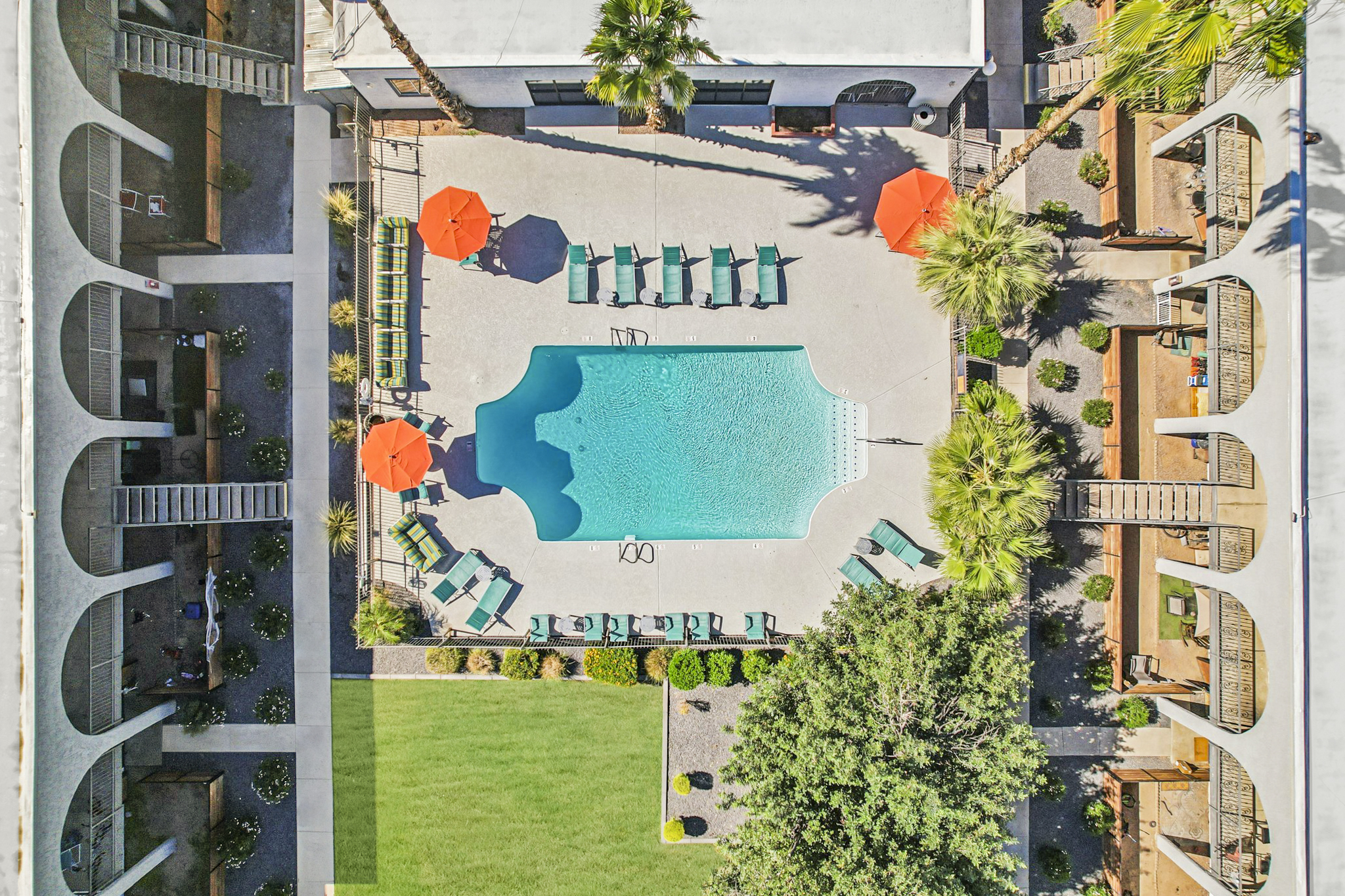 Aerial view of a hotel courtyard featuring a large swimming pool surrounded by lounge chairs and orange umbrellas. Lush greenery and landscaped areas are adjacent to the pool, with balconies visible from surrounding buildings. The scene is bright and inviting, perfect for relaxation.