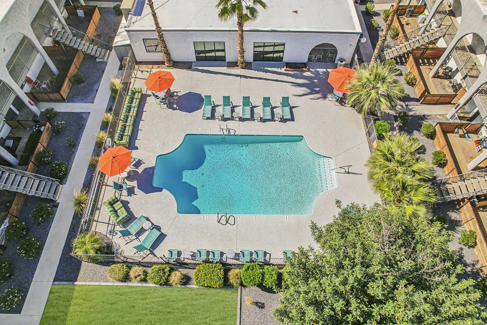 Aerial view of a swimming pool surrounded by lounge chairs and orange umbrellas. Lush greenery and palm trees are visible nearby, along with a well-maintained courtyard area. The layout features a clean and inviting design, perfect for relaxation and leisure.