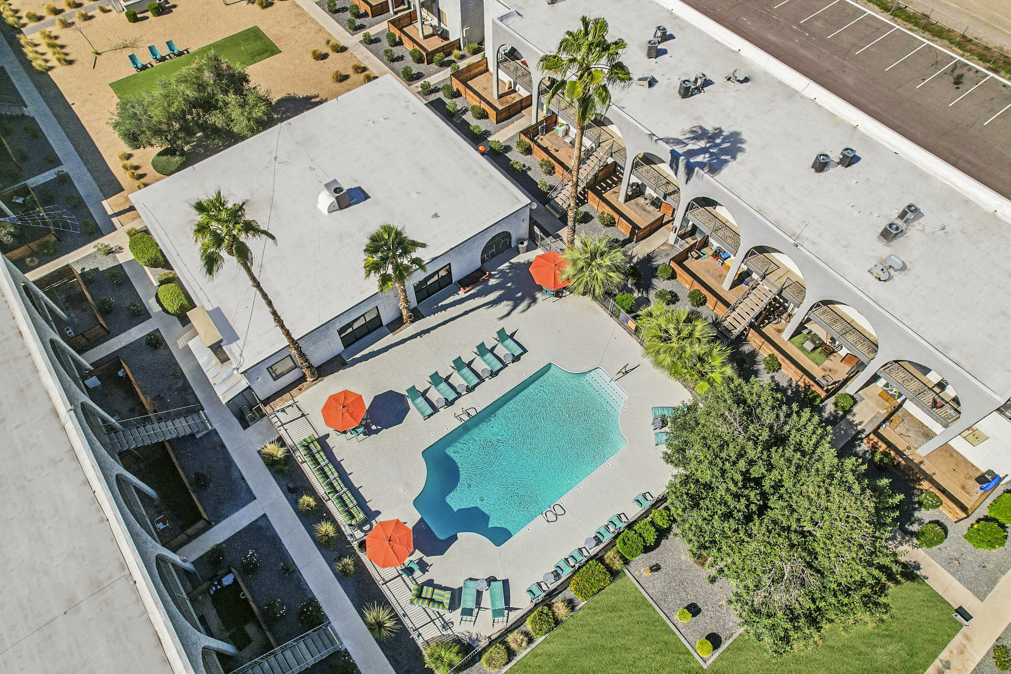 Aerial view of a hotel pool area featuring a large swimming pool surrounded by loungers and orange umbrellas. The landscape includes palm trees and well-maintained greenery, with a building and parking area visible nearby.