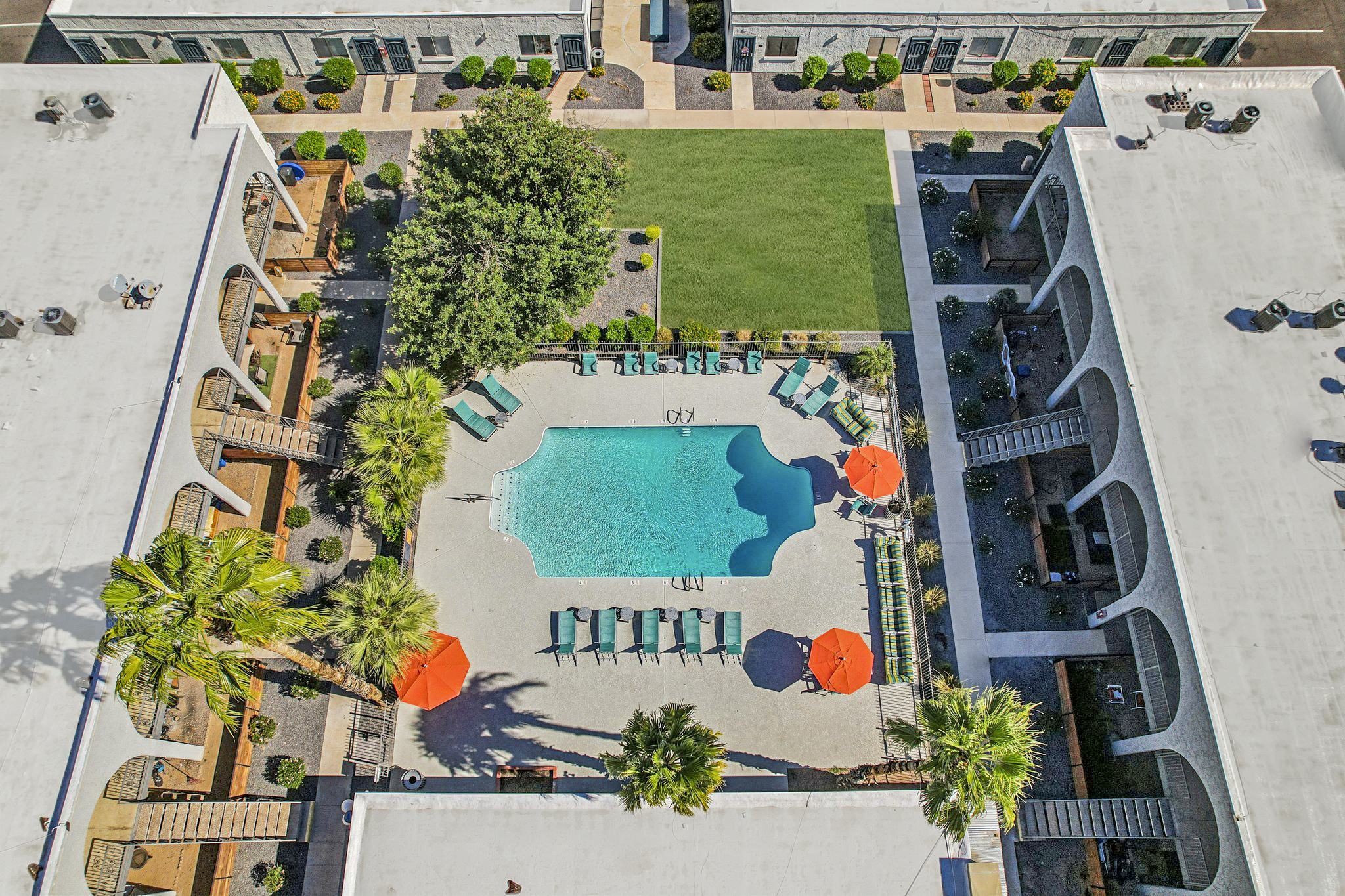 Aerial view of a courtyard featuring a swimming pool surrounded by green lounge chairs and orange umbrellas. The area is landscaped with palm trees and greenery, and there are buildings on either side with patios. The scene is bright and inviting, showcasing a relaxing outdoor space.