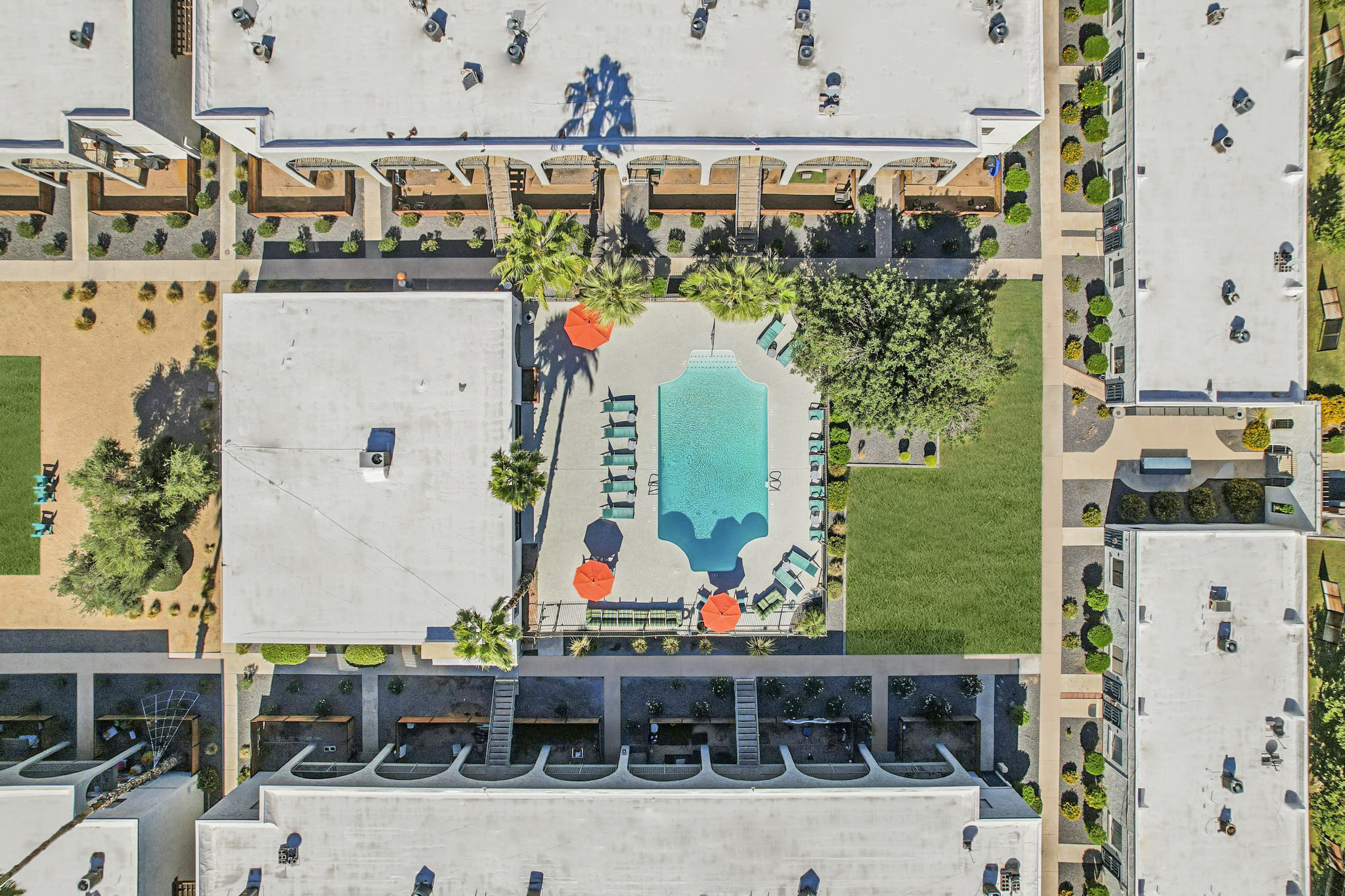 Aerial view of a hotel complex featuring a central swimming pool surrounded by lounge chairs and umbrellas. The area includes landscaped gardens and pathways, with buildings surrounding the pool. Clear skies and well-maintained greenery create a relaxing atmosphere.