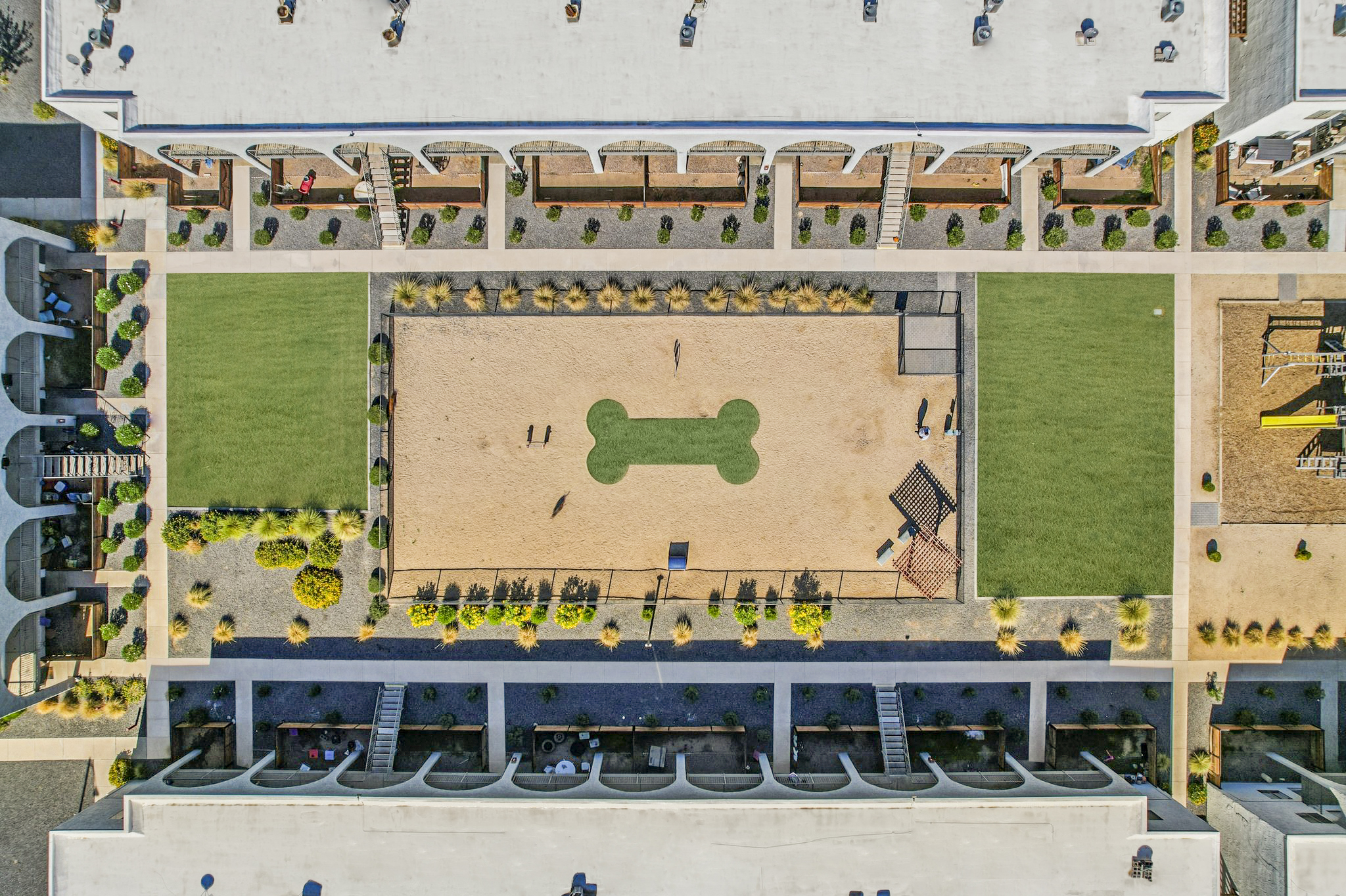 Aerial view of a dog park featuring a large sandy area shaped like a dog bone, surrounded by green grass. Nearby, people can be seen walking or playing with dogs. The view includes buildings and landscaped areas around the park.