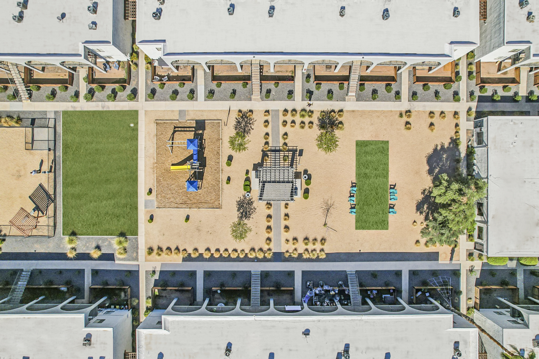 An aerial view of a residential complex featuring landscaped courtyards, including open green spaces, playground equipment, and seating areas. The layout includes various amenities like picnic spots and paths surrounded by buildings.
