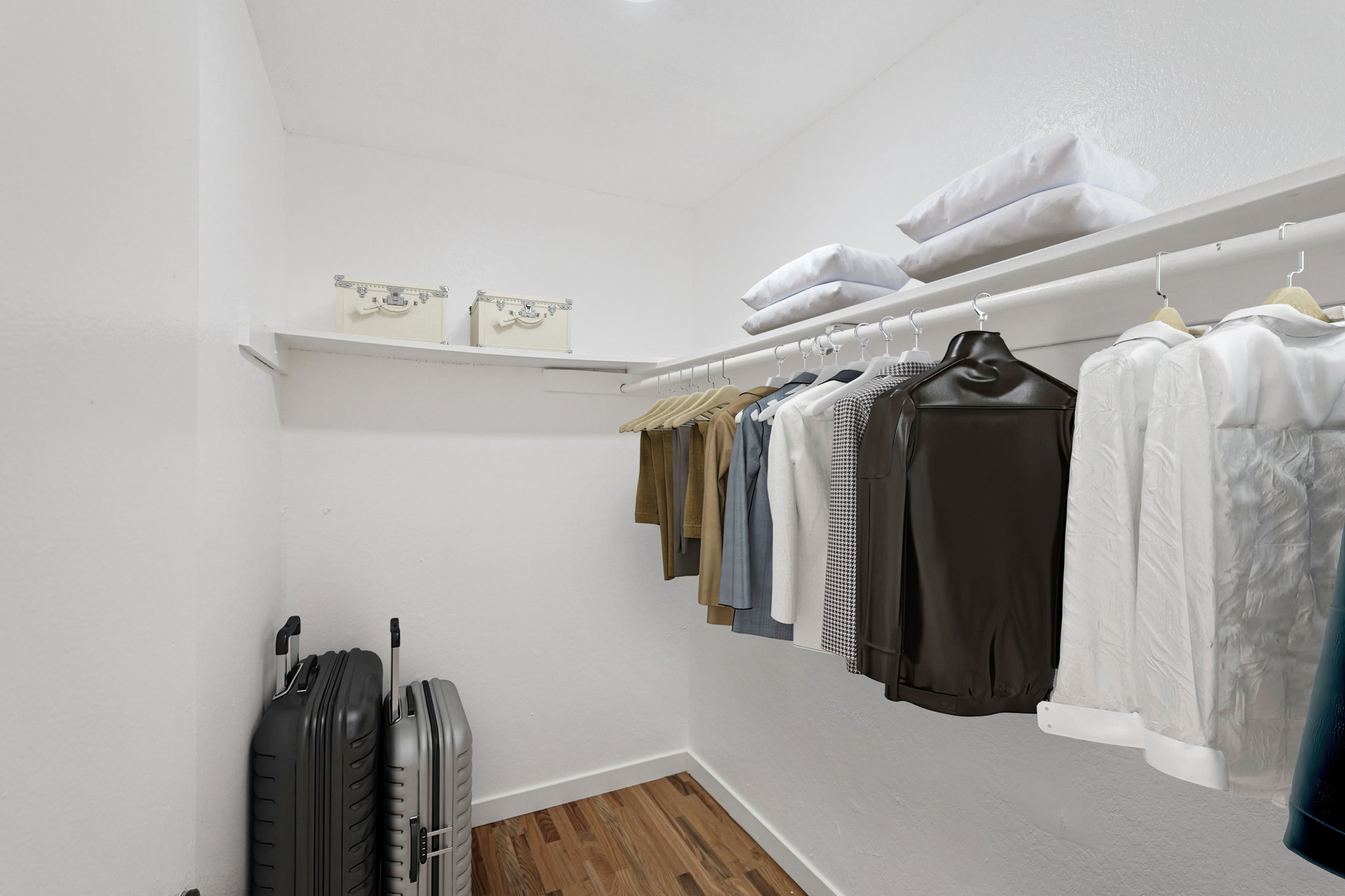 A neatly organized closet featuring a variety of clothing items hanging on hangers, along with two suitcases on the floor and several folded pillows on a shelf. The walls are painted white, and the flooring is wood.