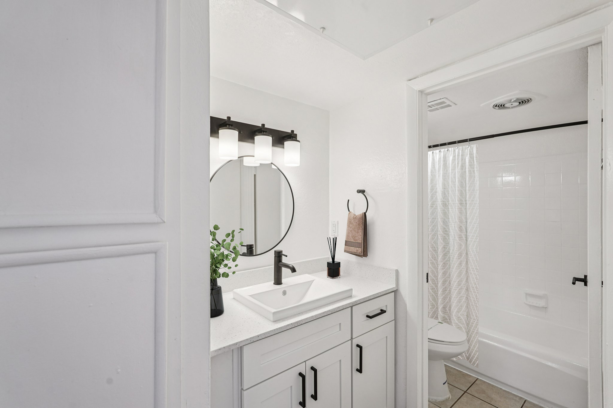 A modern bathroom featuring a circular mirror above a white sink with a stylish black faucet. The countertop is adorned with a small potted plant and a towel. A white shower curtain is visible in the background, along with a toilet and tiled flooring, all in a clean, minimalistic design.