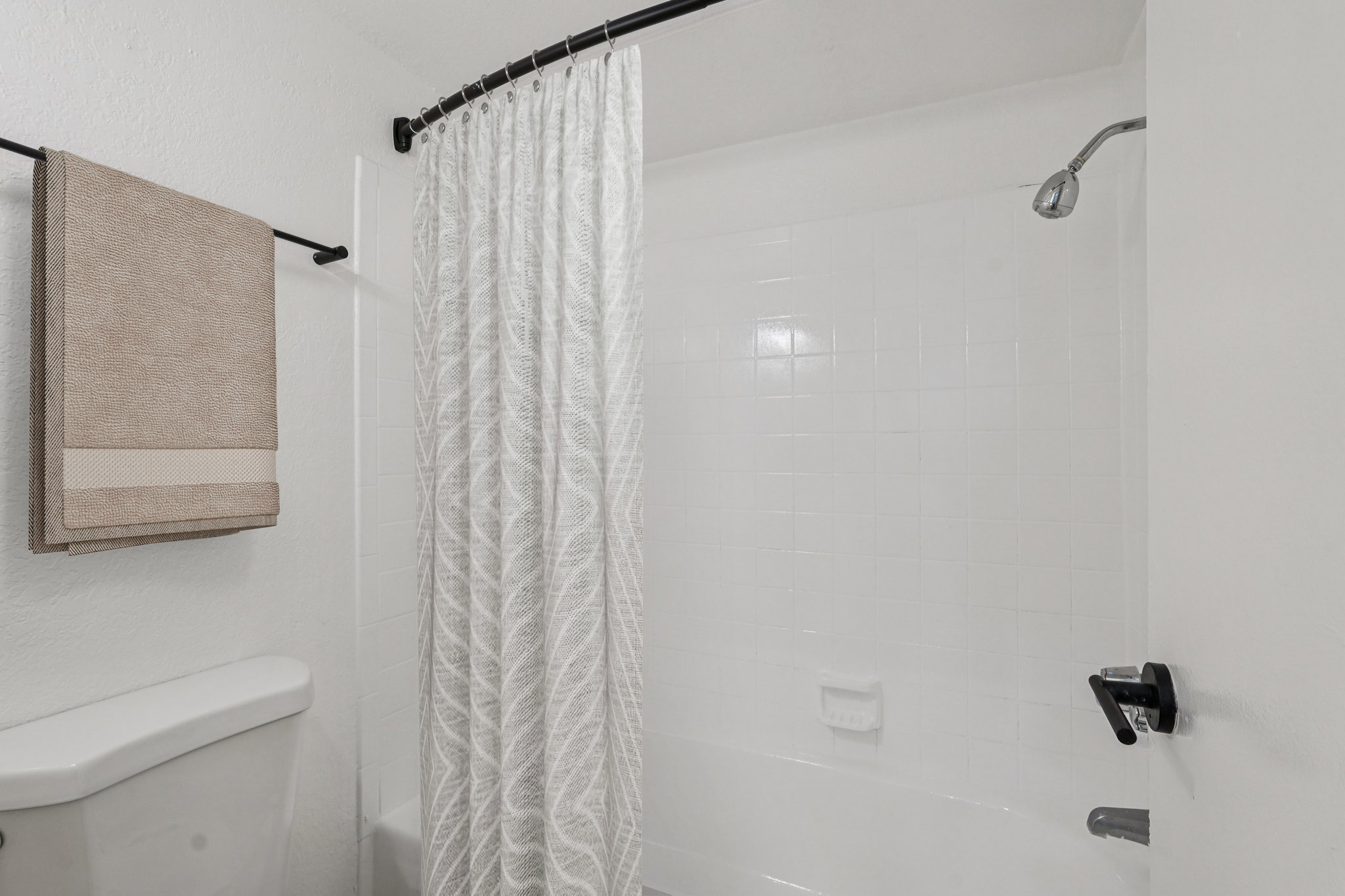 A view of a bathroom shower area featuring a white shower curtain, a chrome showerhead, and a light-colored wall with tiled accents. A neatly folded towel hangs on a towel rack next to the curtain. The overall ambiance is clean and modern.