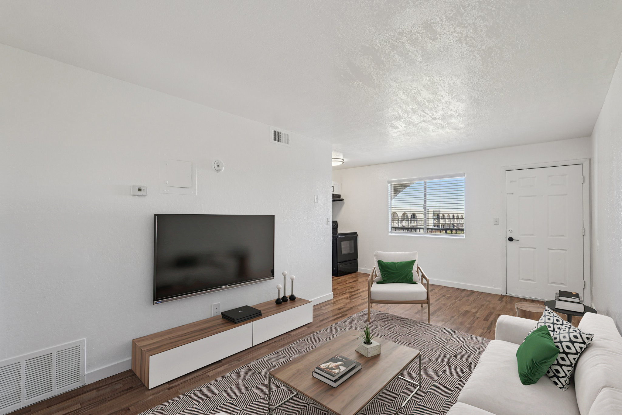 Modern living room featuring a flat-screen TV mounted on the wall above a minimalist white media console. A stylish coffee table with a small plant sits on a patterned rug, and a white couch with green accent pillows is positioned nearby. Natural light filters in through a window, illuminating the space.