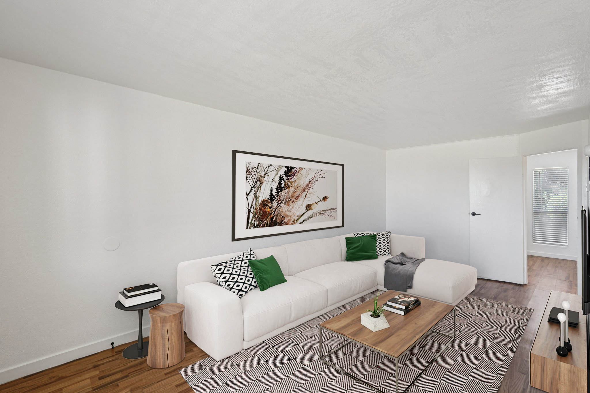 A modern living room featuring a white sectional sofa with green and black accent pillows, a wooden coffee table topped with decorative items, and a framed artwork on the wall. The room has light-colored walls and wooden flooring, with a door and window visible in the background.
