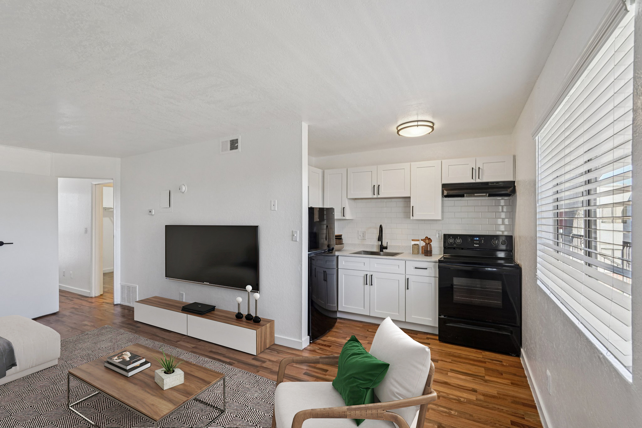A modern, open-concept living area featuring a cozy sofa, a wooden coffee table, and a large TV. The adjacent kitchen has white cabinets, black appliances, and a tiled backsplash. Natural light enters through a window with blinds, and a textured area rug adds warmth to the space.