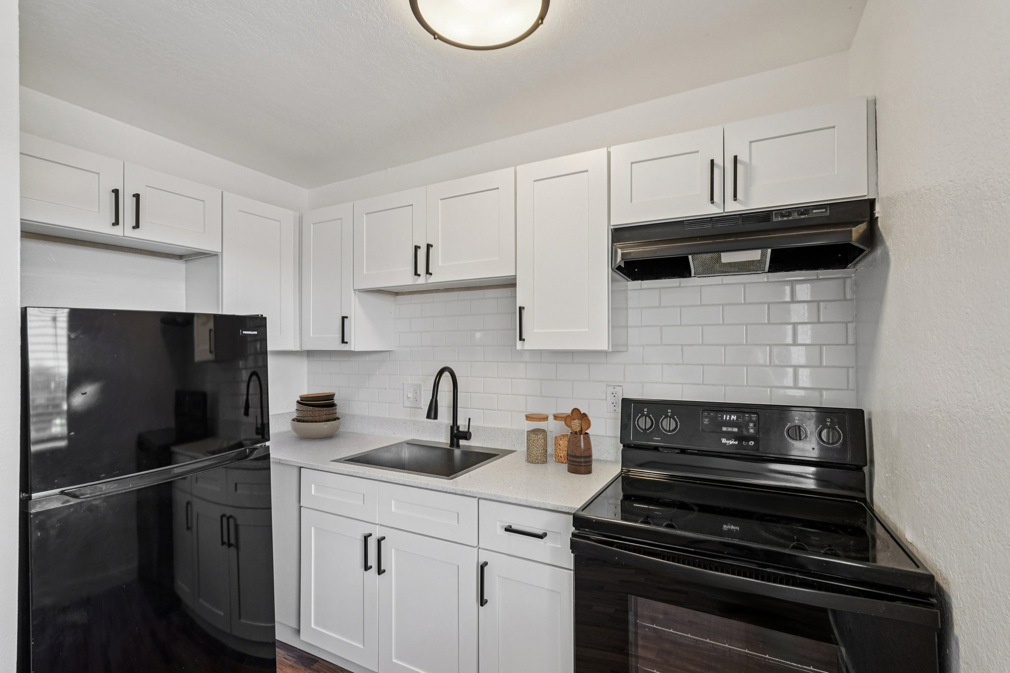 Modern kitchen featuring white cabinets, a black refrigerator, and a black stove. The countertop is light-colored with a sink and decorative items. The backsplash is white subway tile, and there are shelves with bowls and jars. Natural light comes from the ceiling fixture.