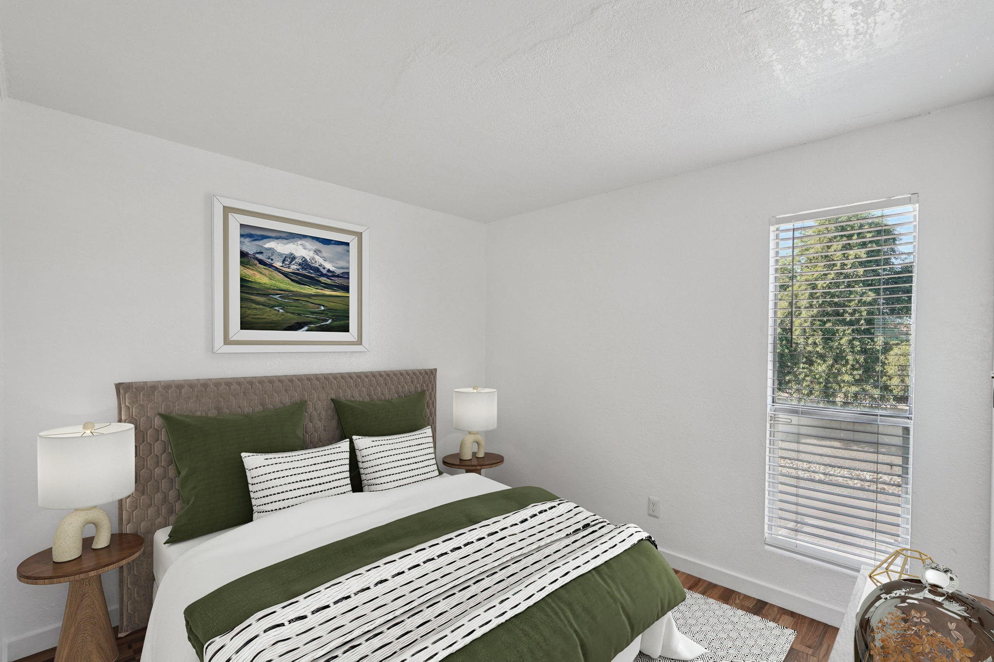 A cozy, well-lit bedroom featuring a queen-sized bed with green and white bedding. Two stylish nightstands with lamps flank the bed. A large window with blinds lets in natural light, and a landscape painting hangs above the headboard. The walls are white, and the floor is wooden, creating a warm atmosphere.