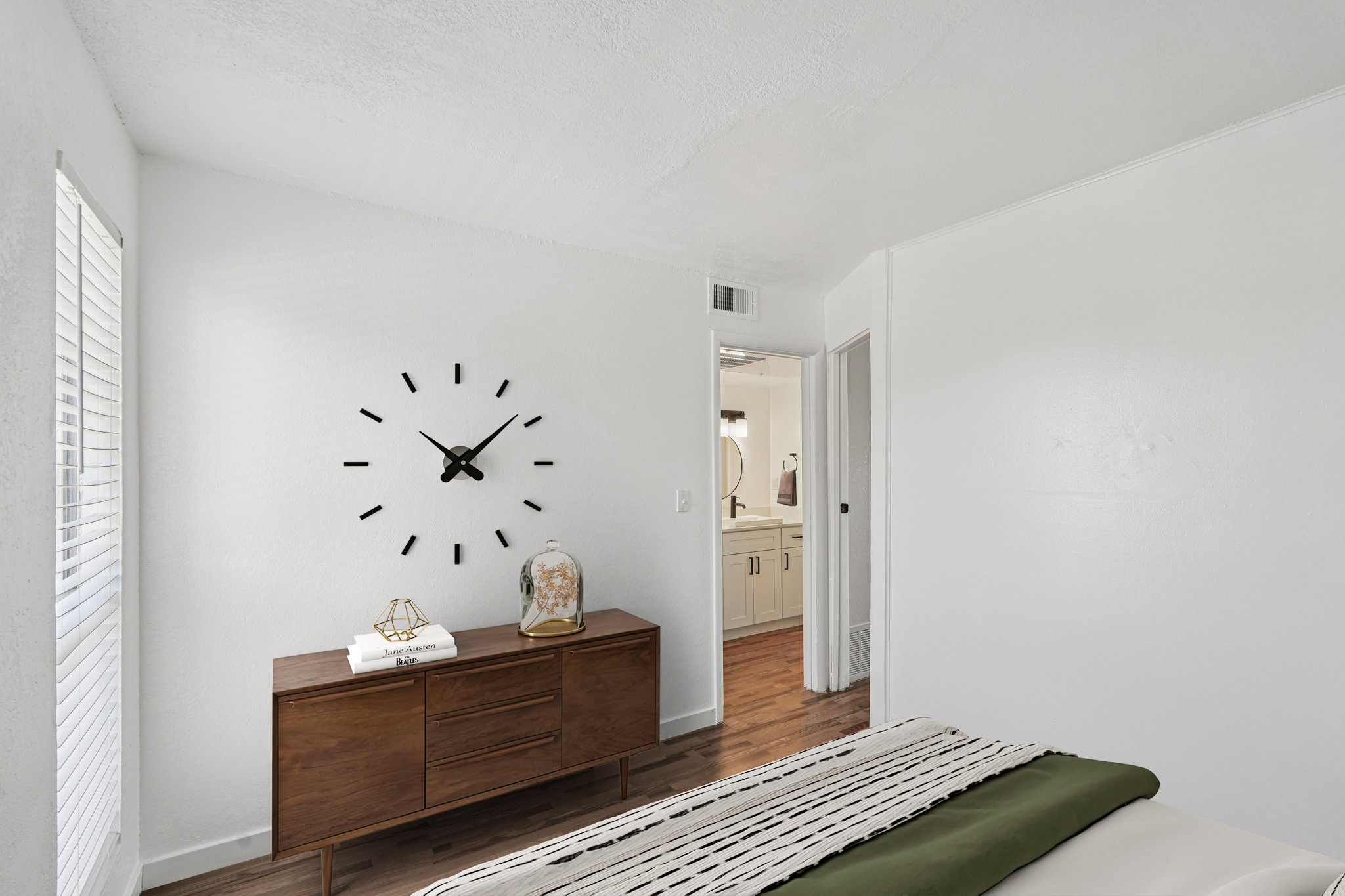 A modern bedroom featuring a white wall and wooden floor. A stylish clock with black hands is mounted on the wall. A wooden dresser sits alongside a bed with a green throw blanket. A doorway leads to a bathroom, visible in the background. The overall decor is minimalistic and contemporary.