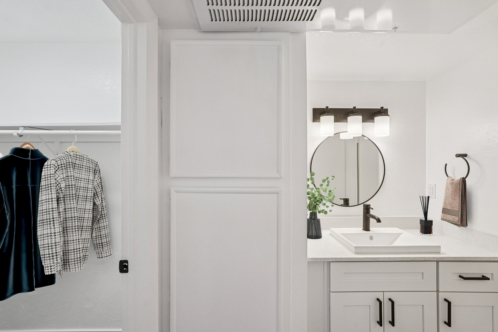 A modern bathroom featuring a circular mirror above a white sink, with a small vase of greenery. To the left, there's a closet with a hanging coat and a shirt. The walls are painted white, and the bathroom has sleek cabinetry and a stylish light fixture above the mirror.