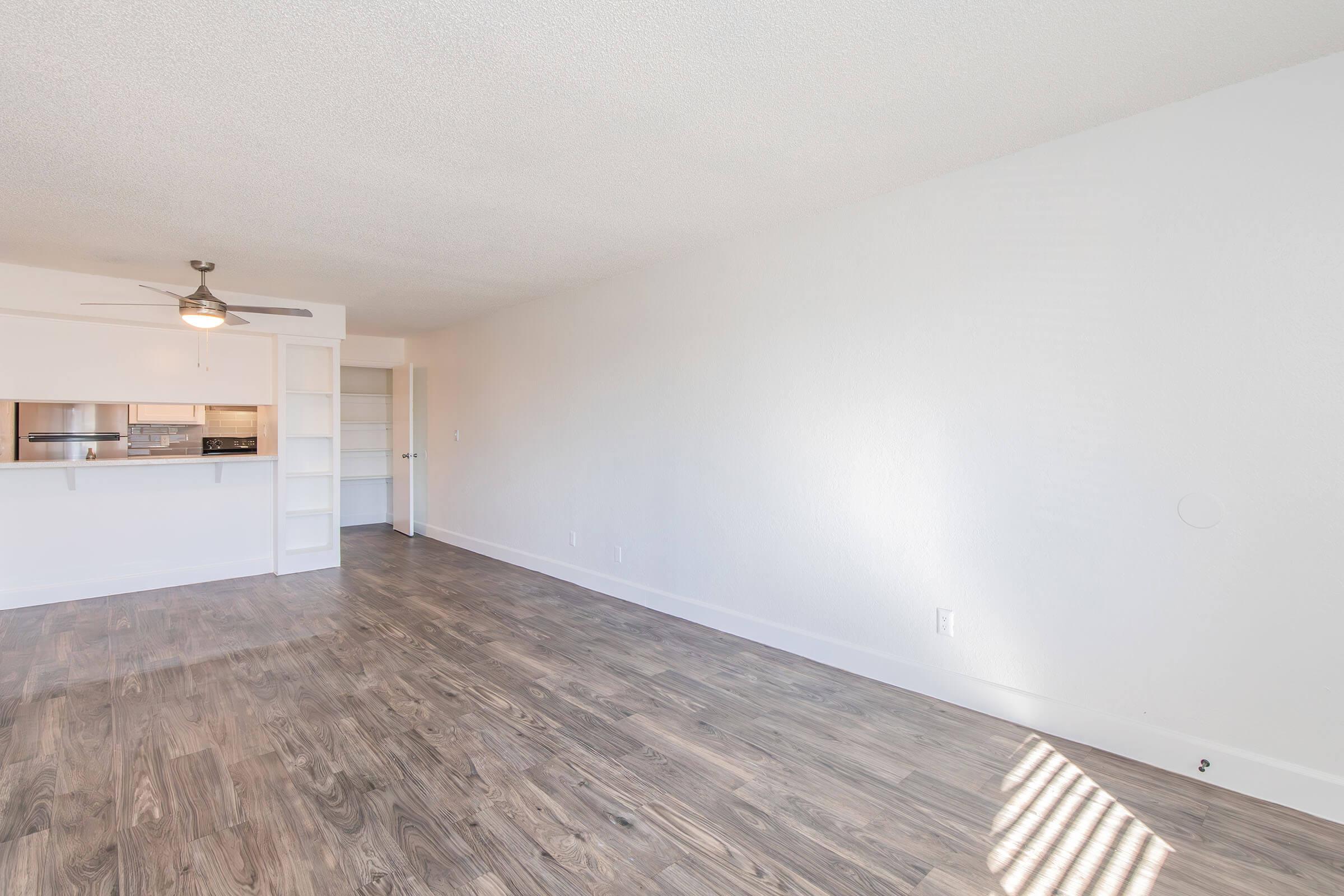 A spacious, empty living room featuring light-colored walls and a ceiling fan. The floor is covered in light wood laminate. In the background, there is an open kitchen area with a bar counter. Natural light streams in through a window on the right, creating a bright and airy atmosphere.
