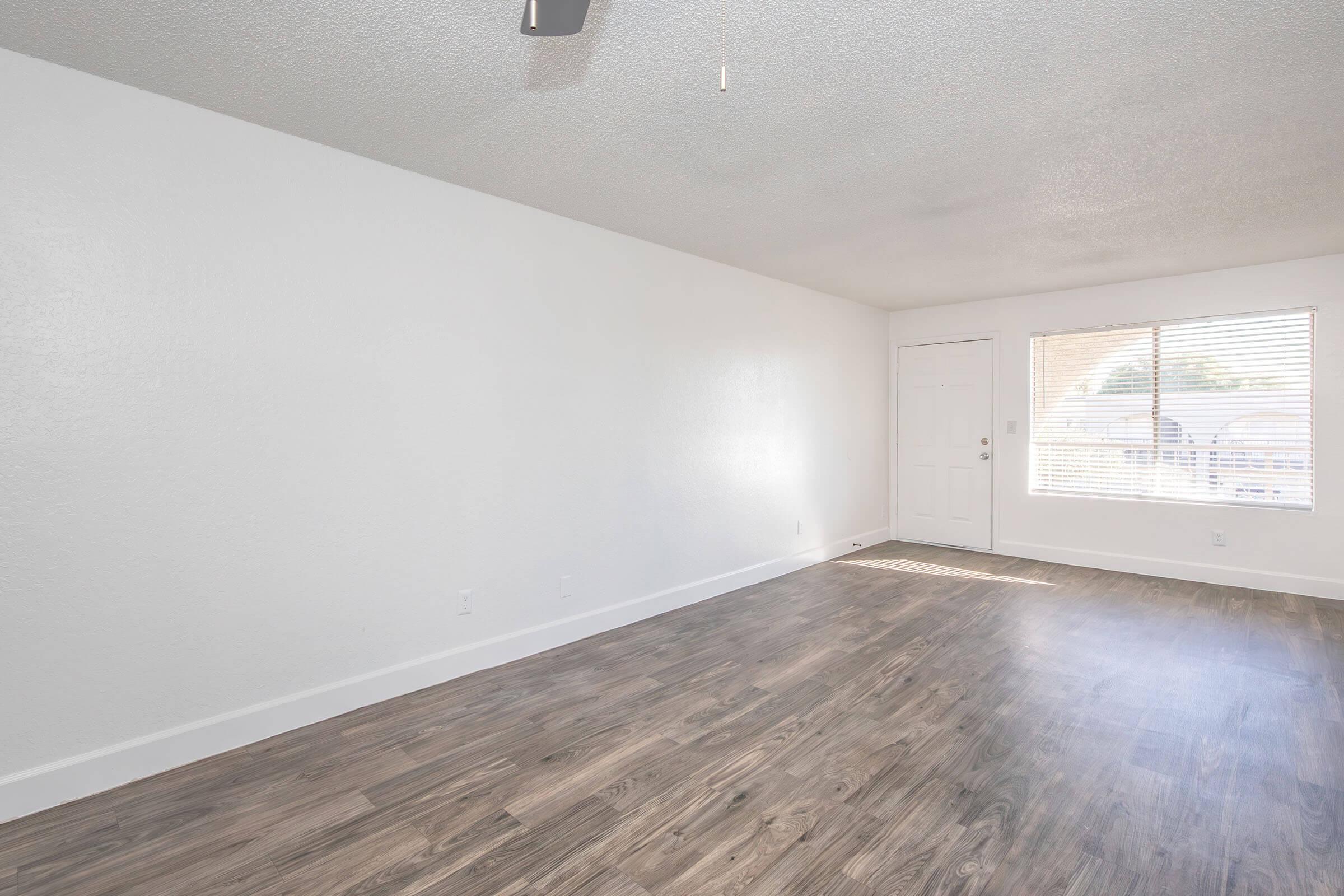 An empty room with light-colored walls and a wood-like floor. There's a window on the right with blinds, allowing natural light to enter. A front door is visible on the left side. The space appears clean and unoccupied, suitable for various uses or decorating styles.