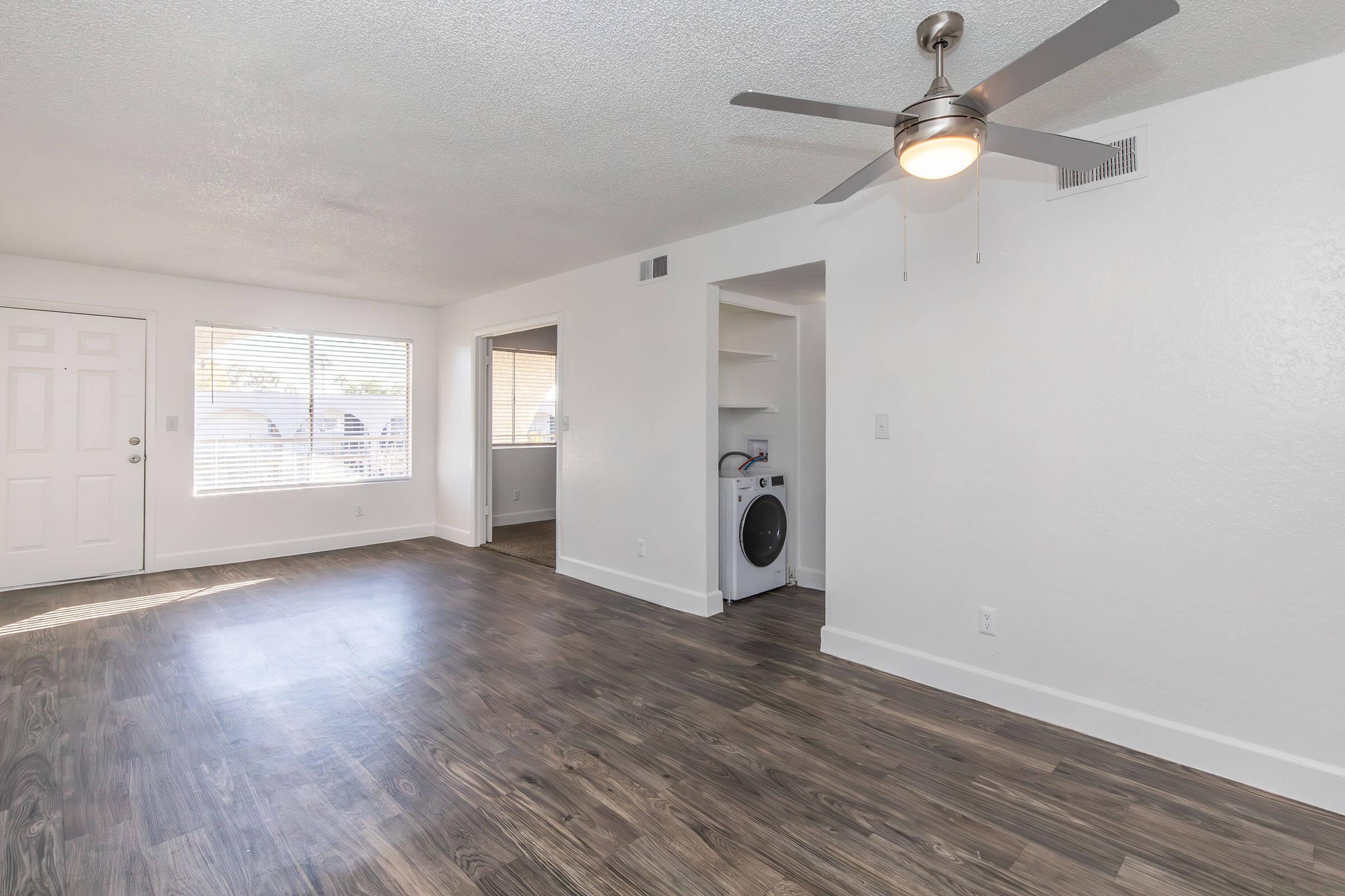 A spacious, well-lit living room featuring light-colored walls, a ceiling fan, and large windows. To the right, there is a laundry area with a washing machine. The flooring is a modern wood laminate, creating a warm and inviting atmosphere.