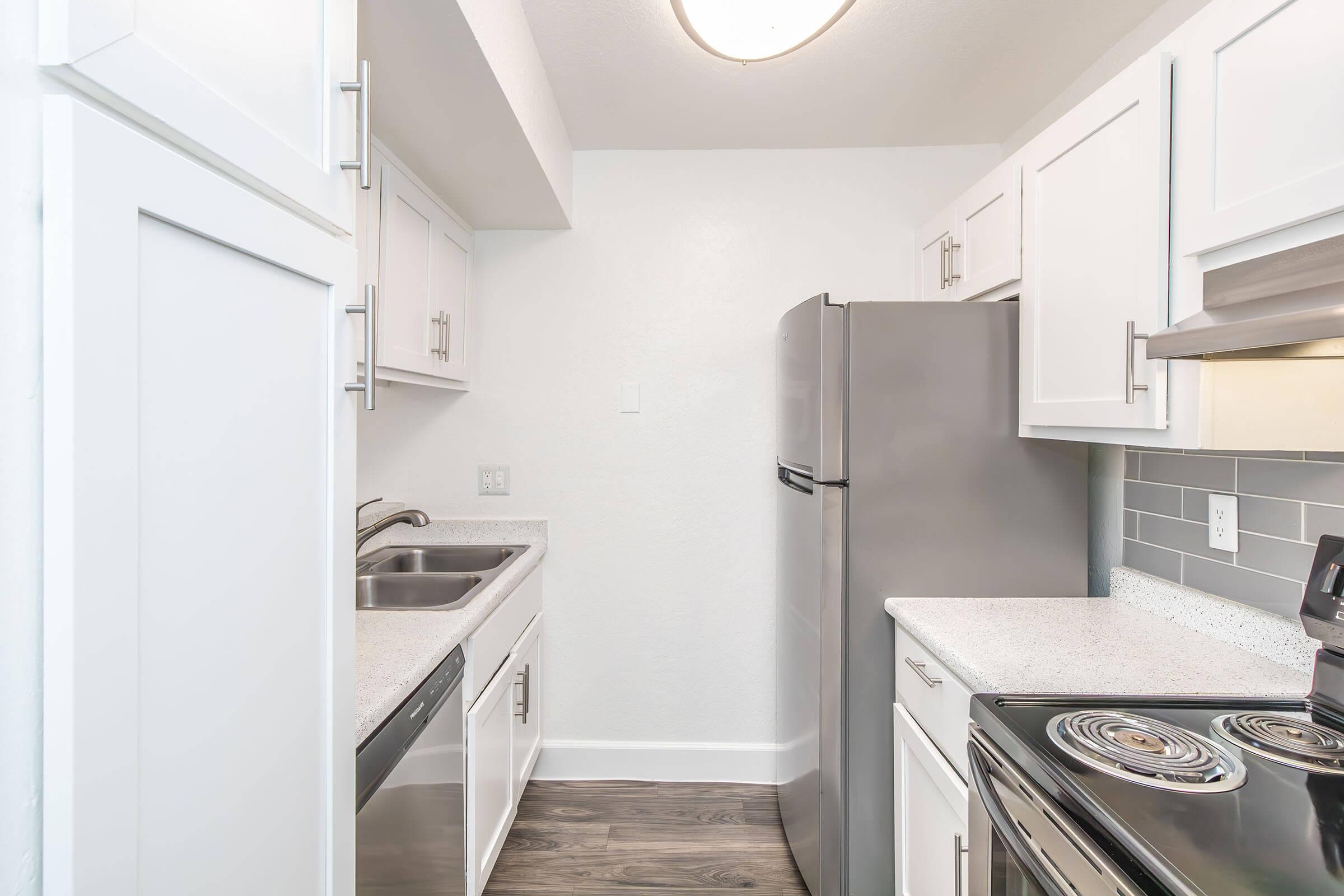 Modern kitchen featuring white cabinets, a stainless steel refrigerator, and an oven. The countertops are light-colored with a speckled pattern, and the walls are painted white. A double sink is installed under a window, and the flooring is a dark wood style. The overall design is clean and contemporary.