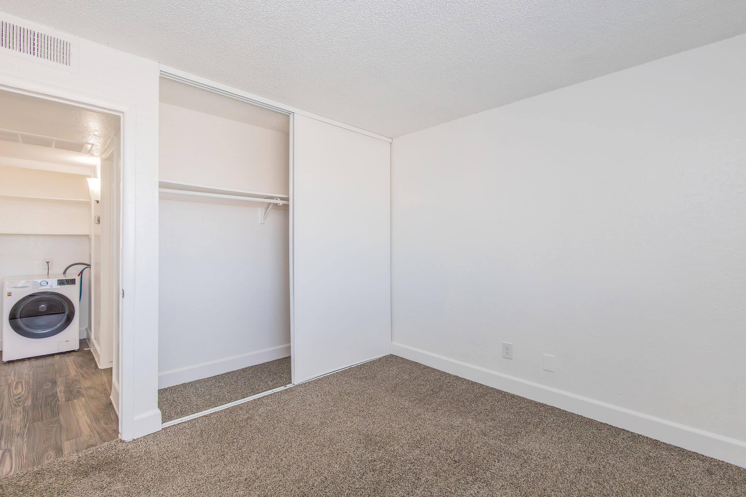 Interior view of a room featuring a wall with a sliding door closet. The closet is empty and there is a small visible area of laundry appliances in the background. The room has beige carpet and white walls, contributing to a bright, clean aesthetic.