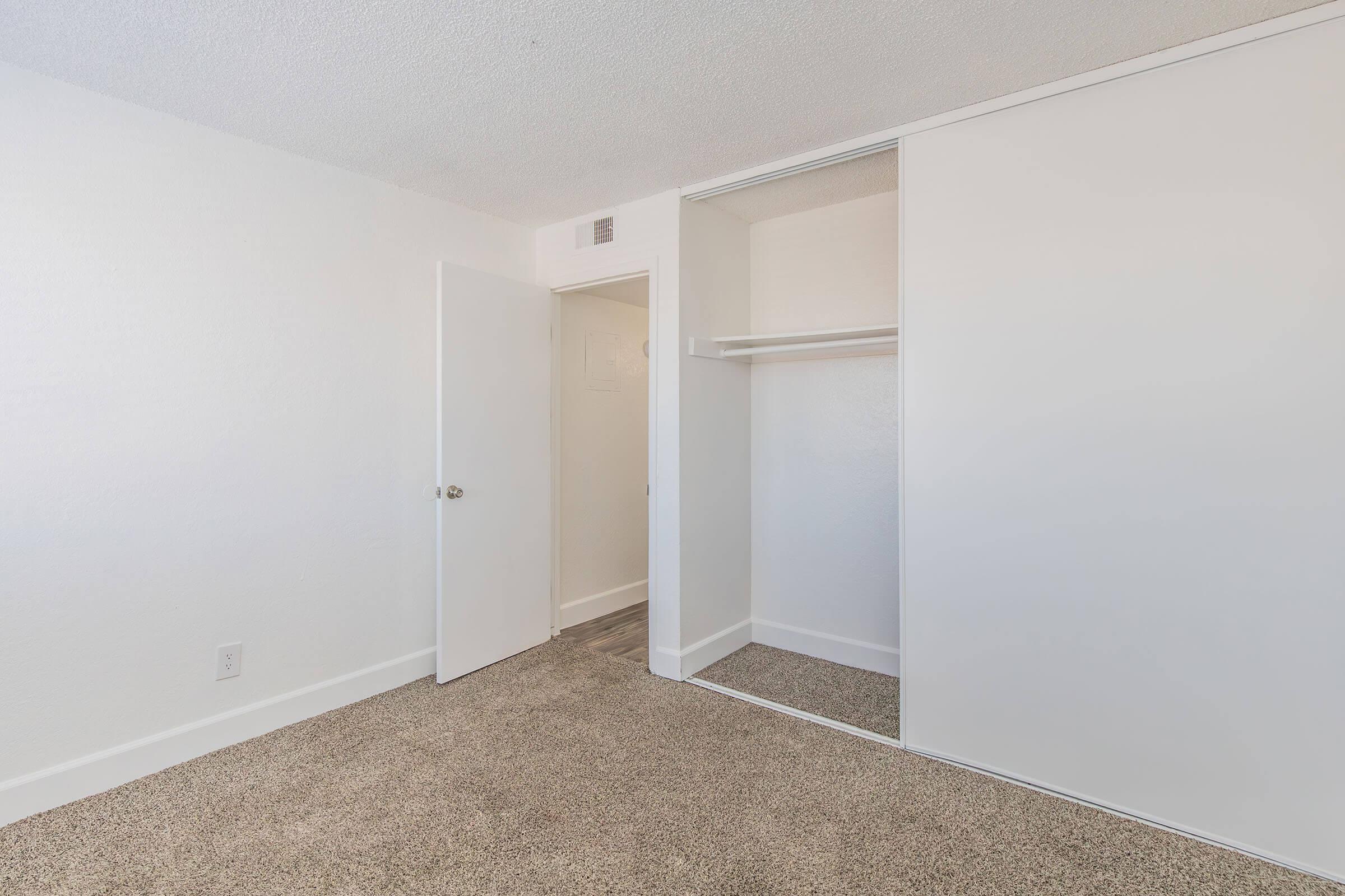 A clean, empty room with light-colored walls and beige carpet. On the left, there is a partially open door leading to another space. On the right, a built-in closet with sliding doors is visible, featuring a shelf inside. The overall atmosphere is bright and uncluttered, suggesting a fresh space ready for furnishing.
