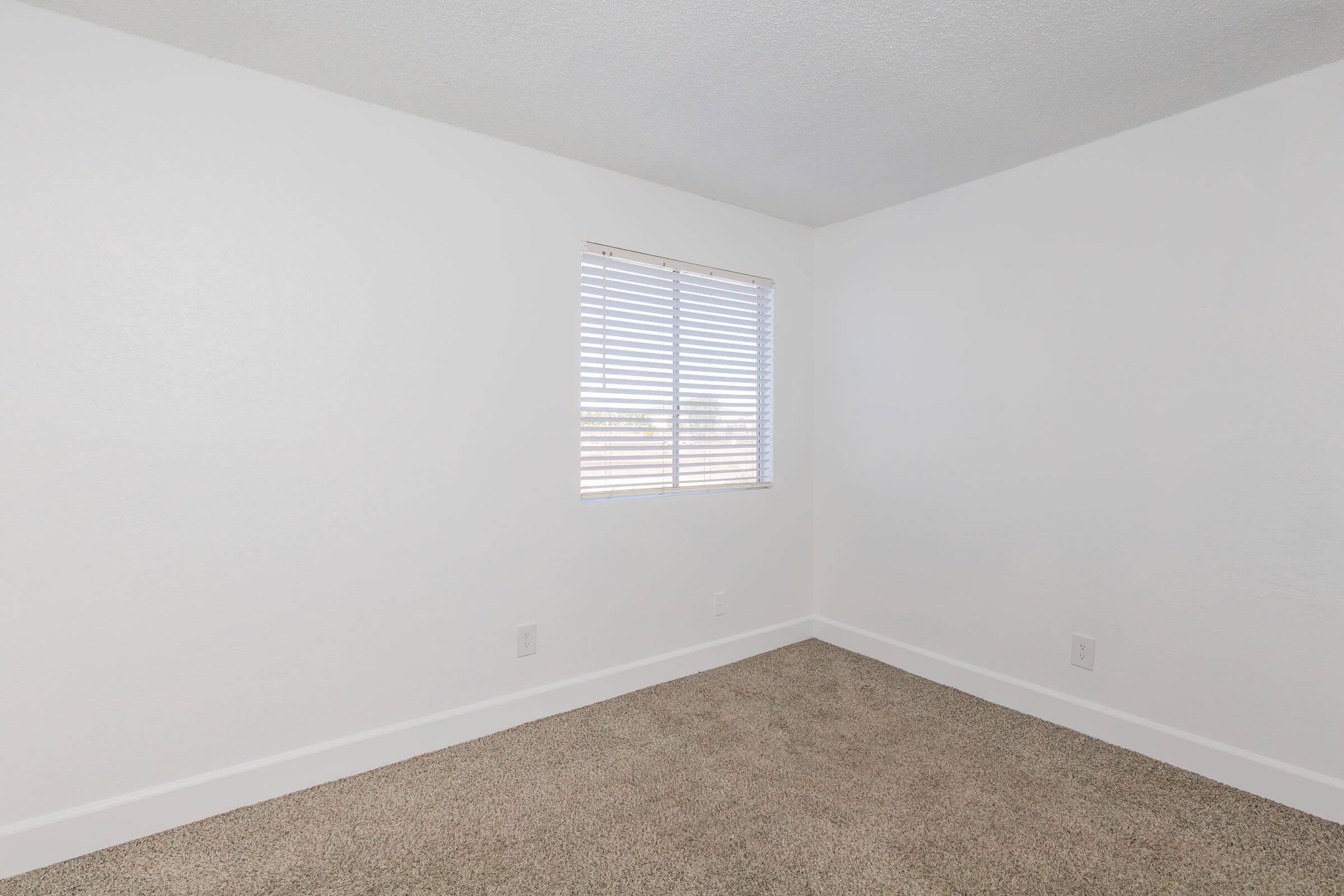 A small, empty room with light beige carpet and white walls. There is a window with white blinds on one side, allowing natural light to enter. The room is unfurnished and has a clean, minimalist appearance.