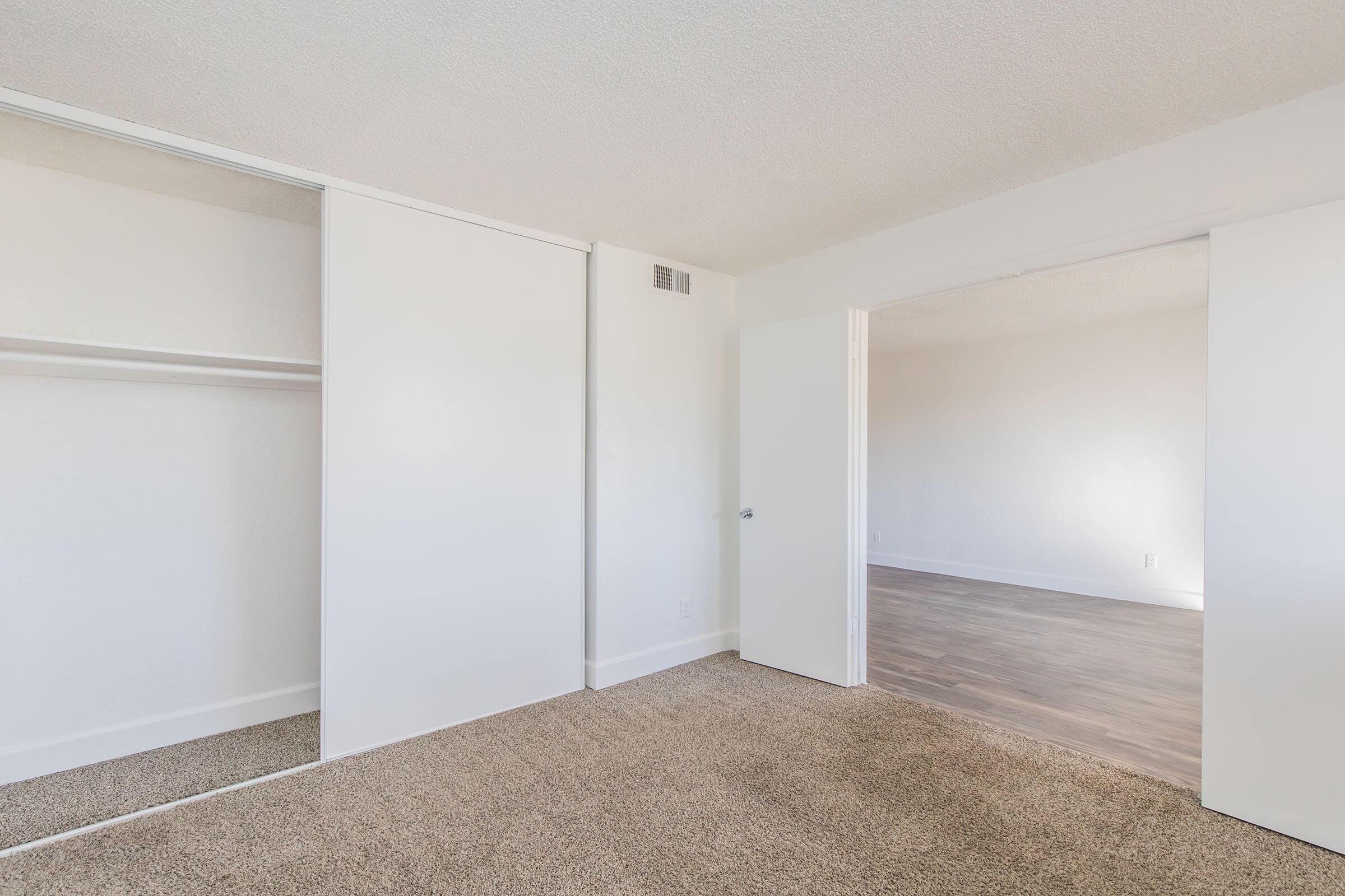A spacious, empty room with beige carpet flooring. One side features a large closet with sliding doors, while the other side has an open doorway leading to another area. The walls are painted white, creating a bright and minimalistic atmosphere.