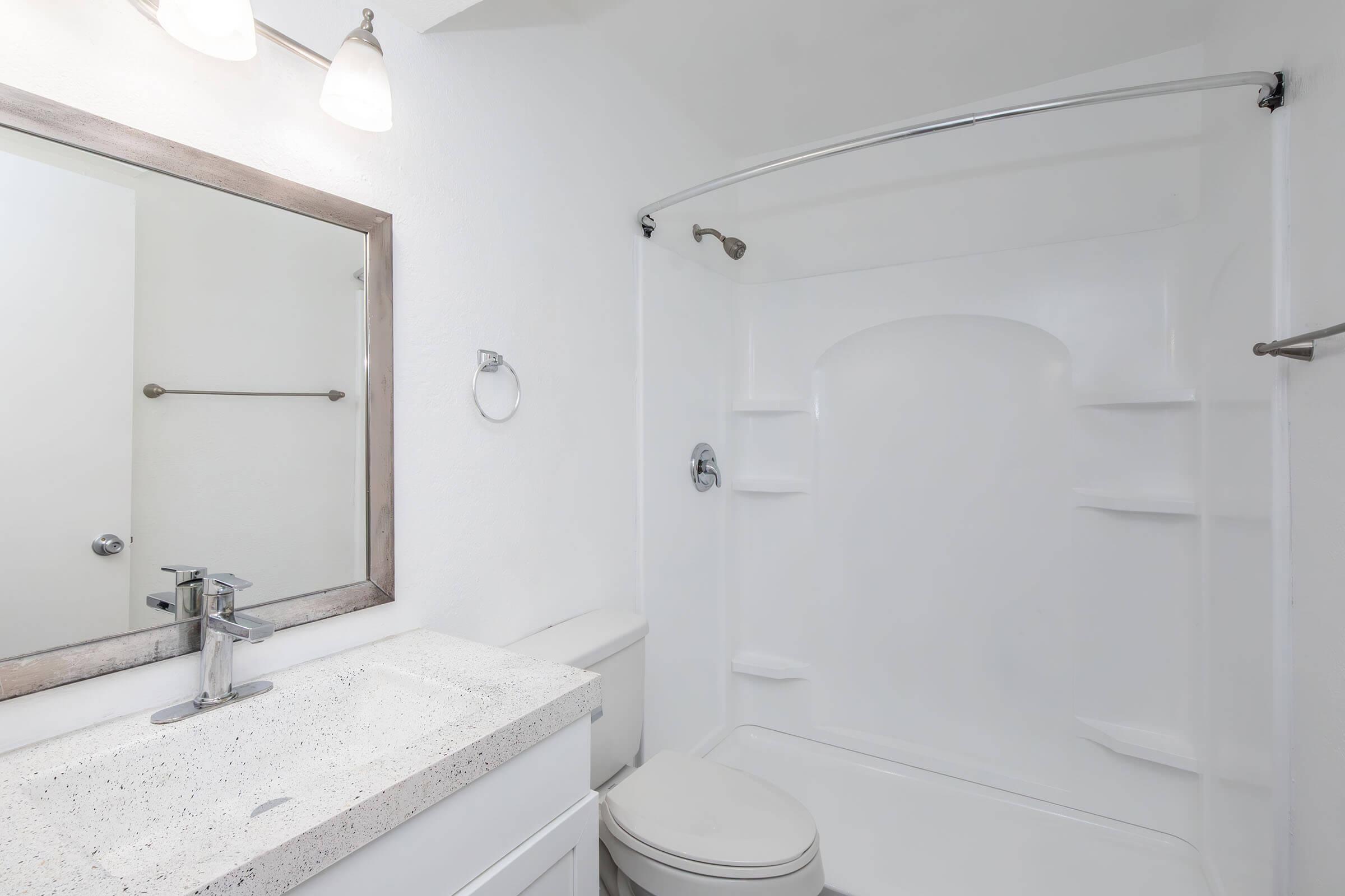 A clean, modern bathroom featuring a white shower enclosure, a sleek toilet, and a vanity with a speckled countertop. The mirror above the sink is framed in wood, and there are two light fixtures above. The walls are painted white, creating a spacious and bright atmosphere.