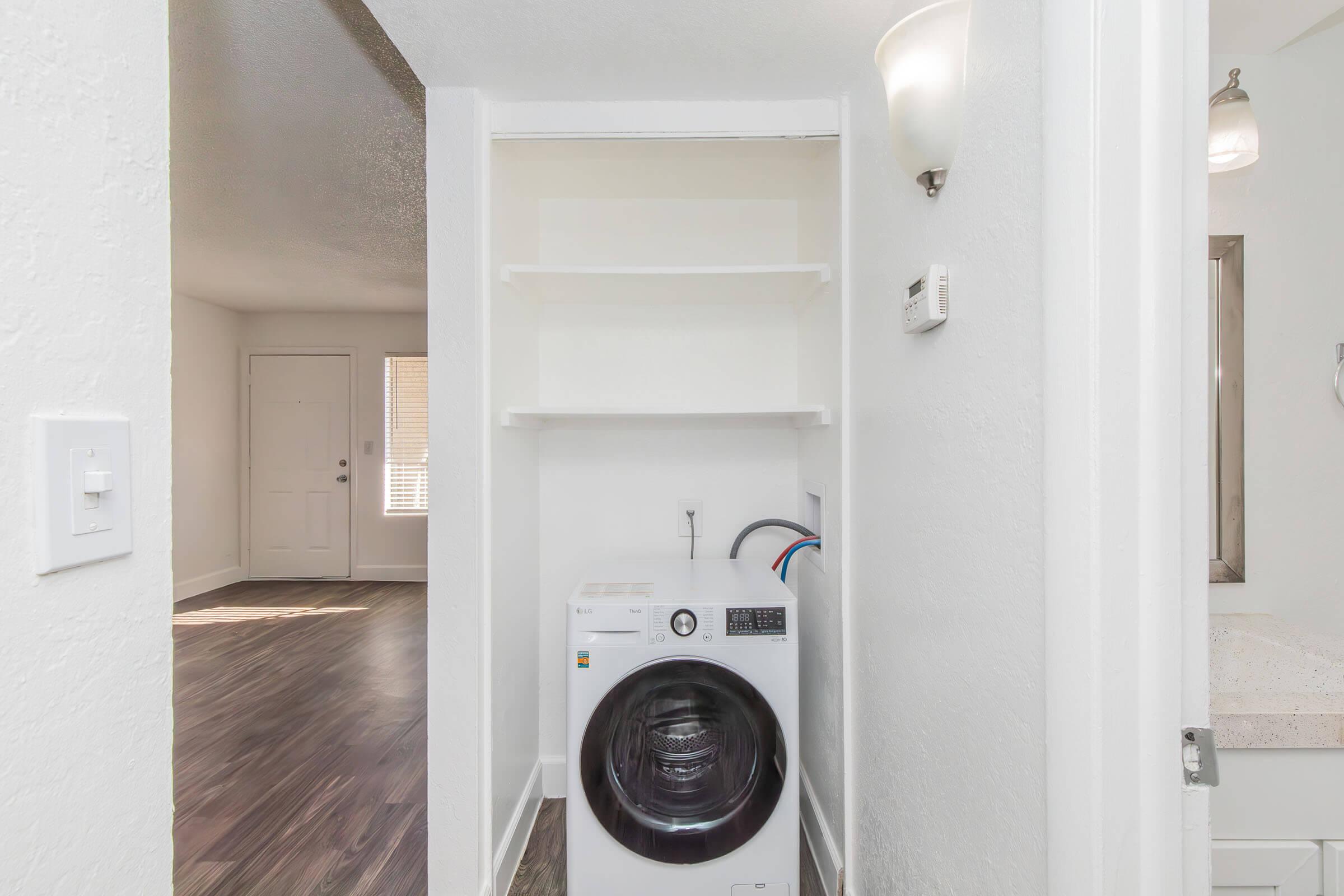 A compact laundry area featuring a front-loading washing machine set against a neutral wall. Above, there are open shelves for storage. The room extends to a doorway leading outside, with natural light coming in through adjacent windows. Stylish lighting fixtures add to the ambiance.