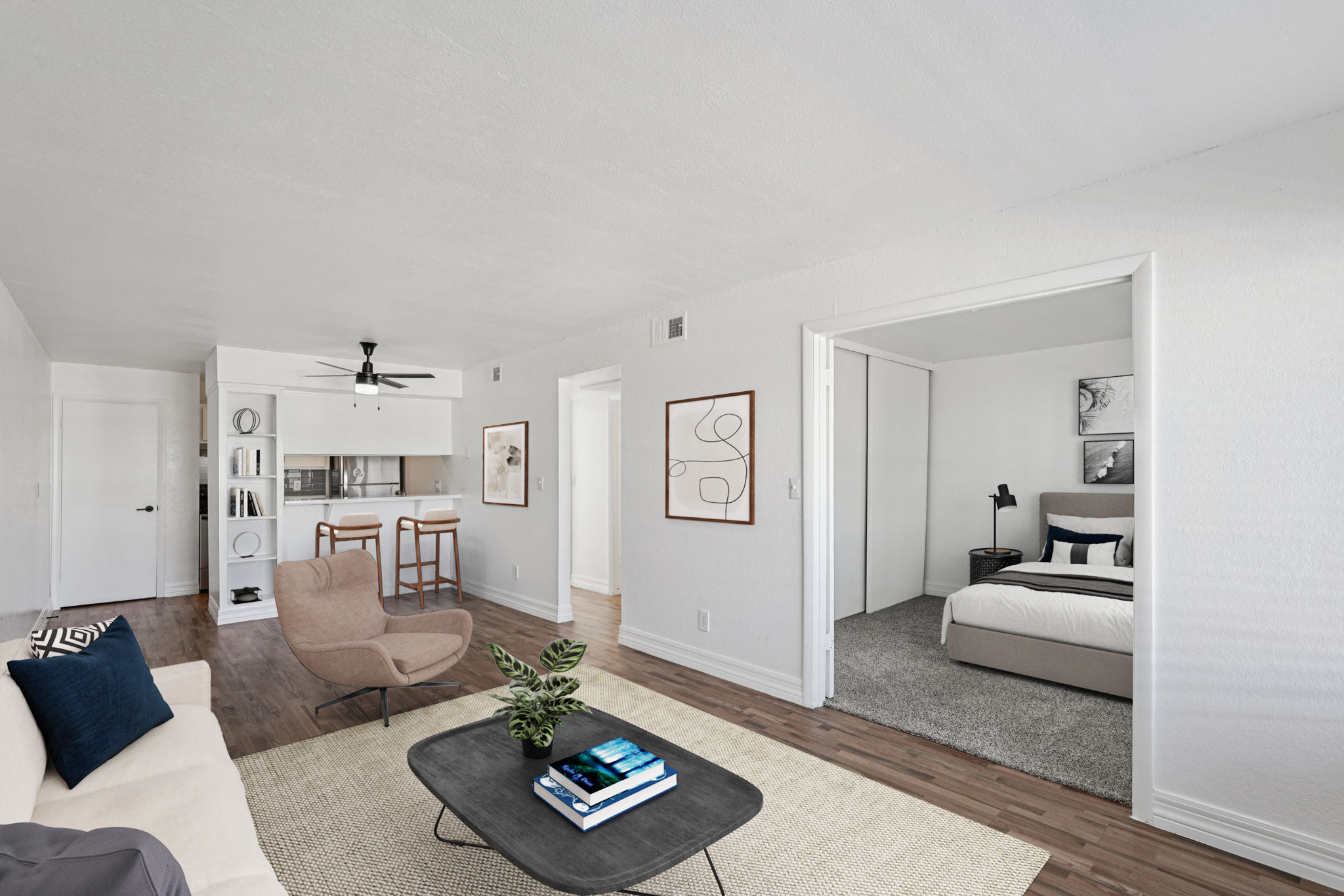 A modern living room featuring a light-colored sofa and a round coffee table with a plant. To the right, a doorway leads to a cozy bedroom with a bed and bedside lamp. The open layout includes a kitchen area visible in the background, accented by neutral tones and minimalistic decor.