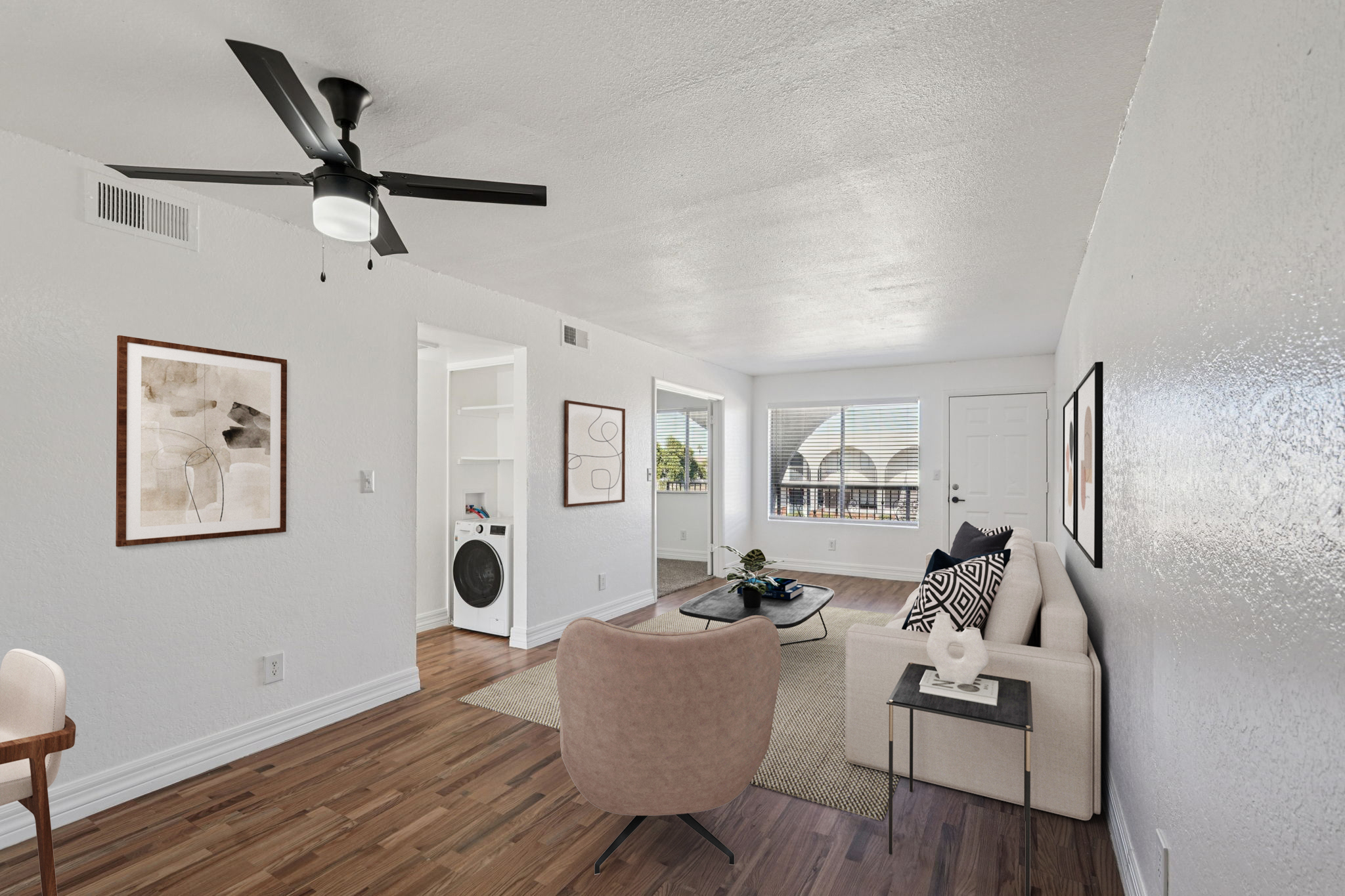 A modern living room featuring a light color palette, hardwood flooring, and a ceiling fan. Furnished with a beige sofa and stylish armchair, the space includes a coffee table with decorative items. Large windows provide natural light, and a white washing machine is visible in a nearby alcove.