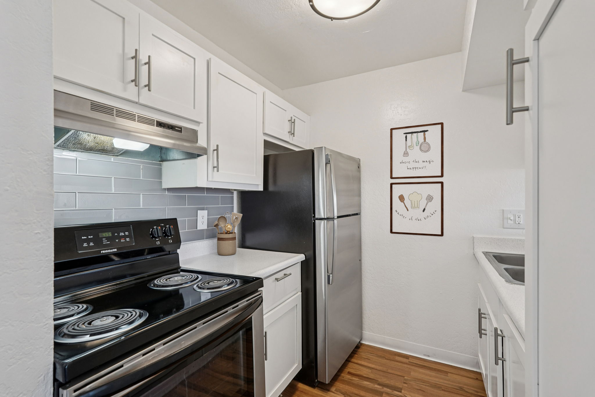 Modern kitchen featuring white cabinets, stainless steel appliances, and a sleek countertop. The black oven and stovetop are visible, along with a dishwasher and sink. Wall décor includes framed kitchen utensils for added charm. Natural light enhances the cozy atmosphere.