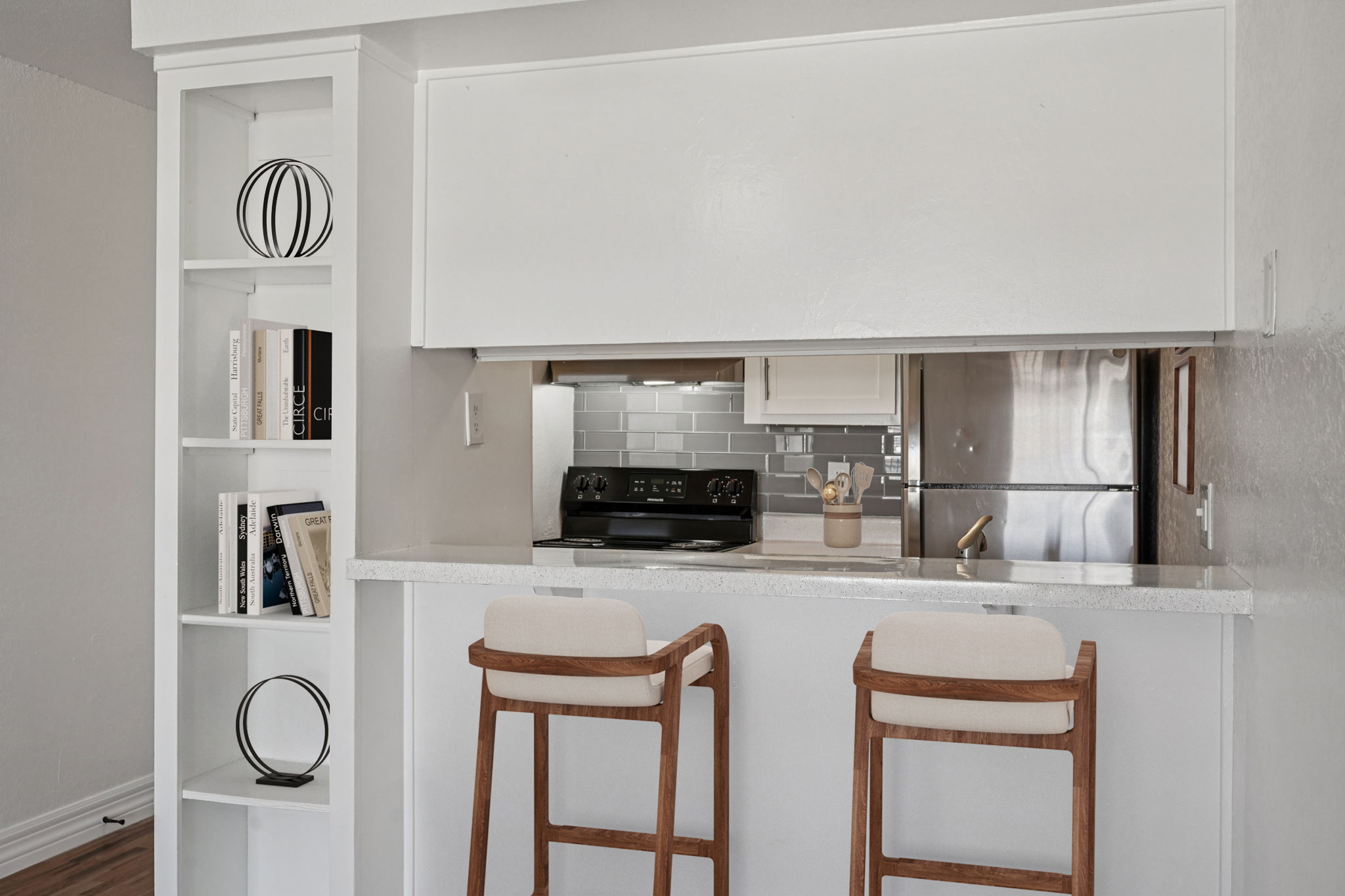 Modern kitchen with a breakfast bar featuring two wooden stools. Open layout showcasing white cabinetry, a glass tile backsplash, and stainless steel appliances. Decorative elements include books on a shelf and a round sculpture. Bright and airy atmosphere with neutral tones.