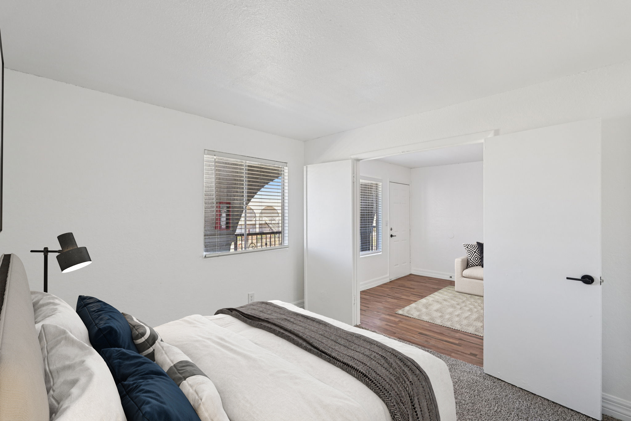 Bright and airy bedroom featuring a neatly made bed with gray and blue bedding. A bedside lamp stands beside the bed. The room includes a window with blinds, allowing natural light, and an open door leading to a living area with a light-colored rug and a couch. The walls are painted white, giving a clean look.